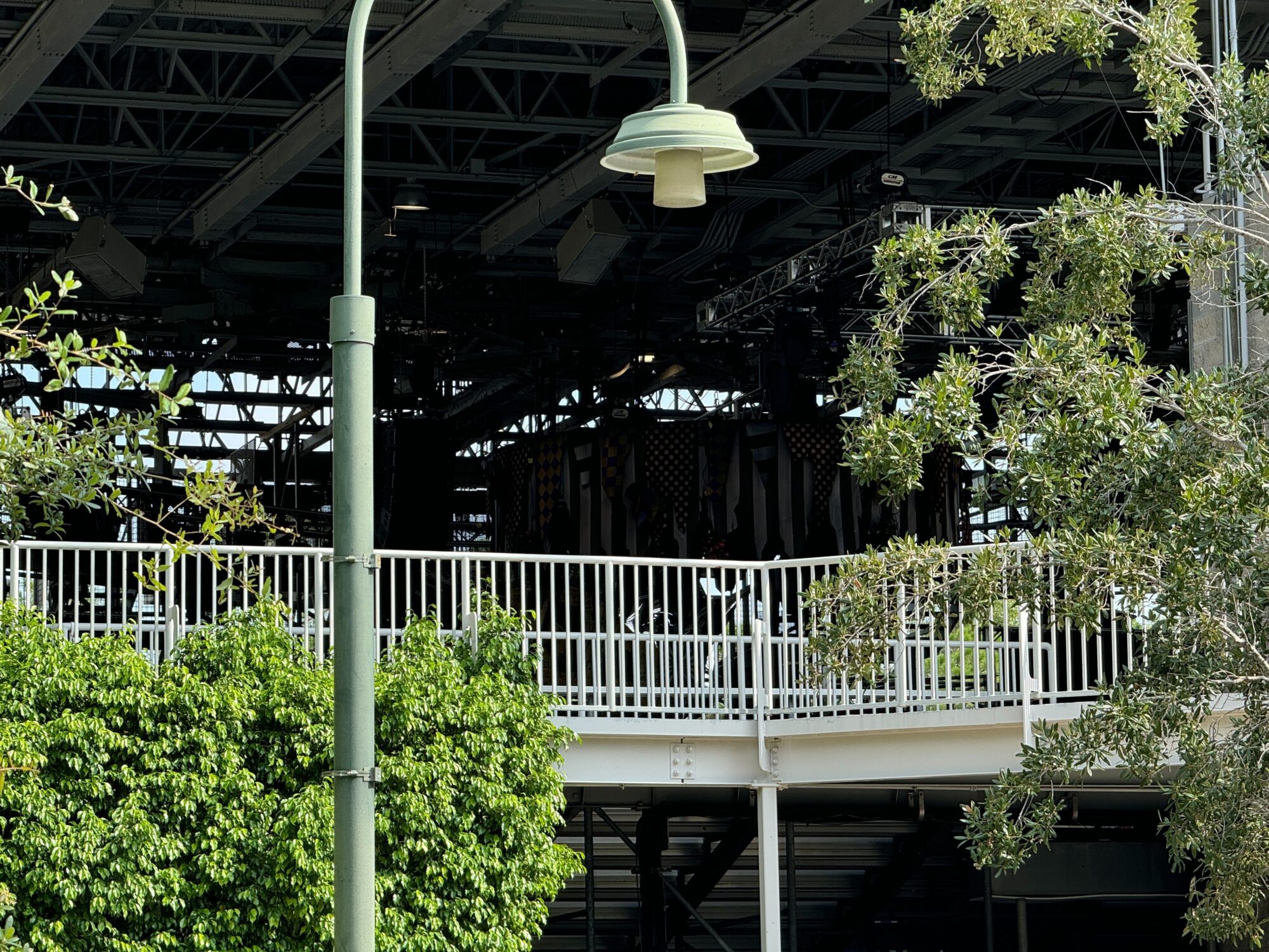 A large stage with lighting and sound equipment is under a metal roof structure. Trees and a railing create a natural barrier in the foreground, framing what looks like an eerie scare zone, with a street lamp casting spooky shadows in front.