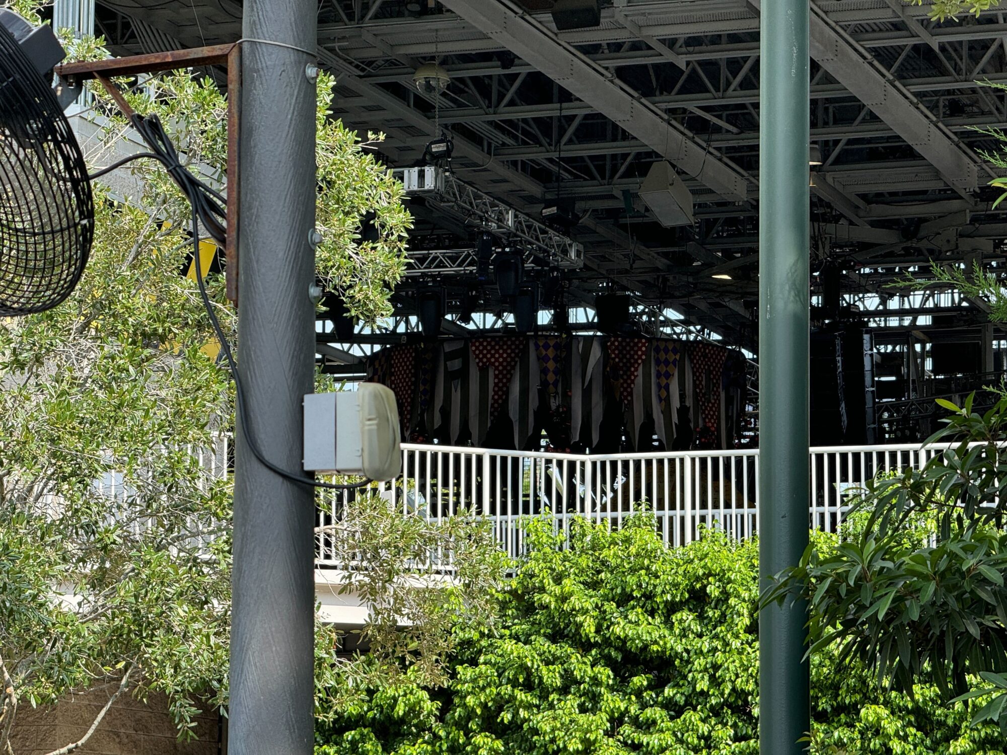 Image of a covered outdoor stage with lighting equipment and a dark-colored backdrop, partially obscured by trees and metal structures, giving it the eerie feel of a scare zone.