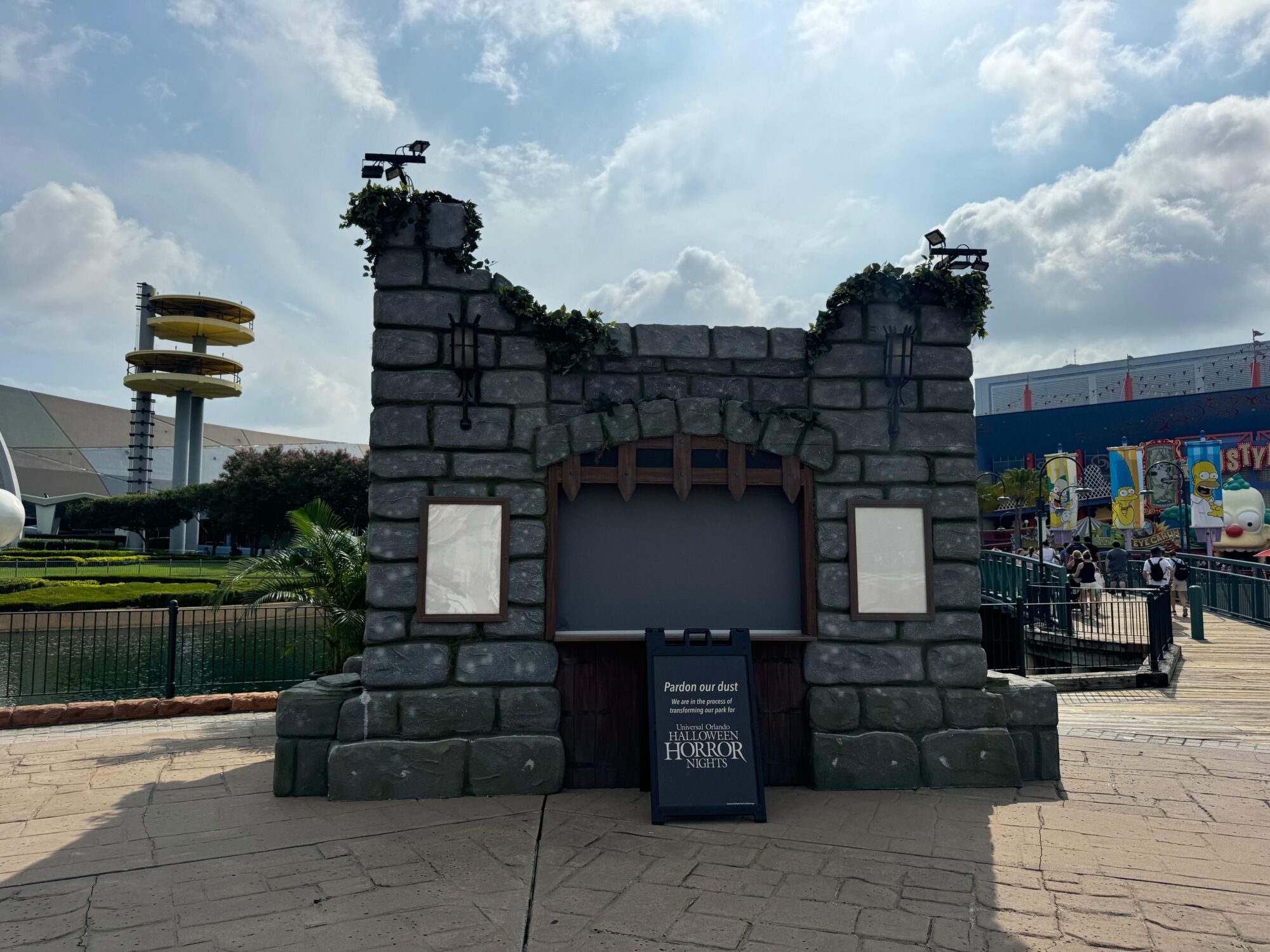 A stone-themed, empty display booth with a "Pardon our dust" sign stands outdoors in a theme park's scare zone, with other attractions and cloudy skies in the background.