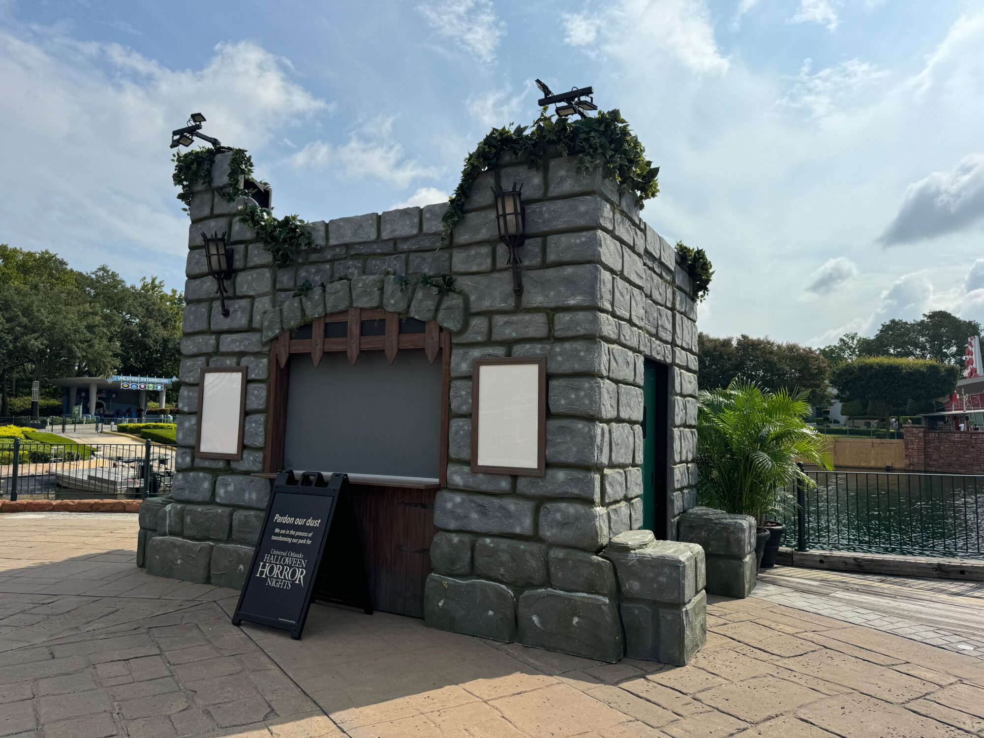 A stone booth resembling a castle features vines, lanterns, and eagle statues. Two menu boards are displayed on the front. A sign at the bottom advertises a nearby harbor marketplace in the scare zone.