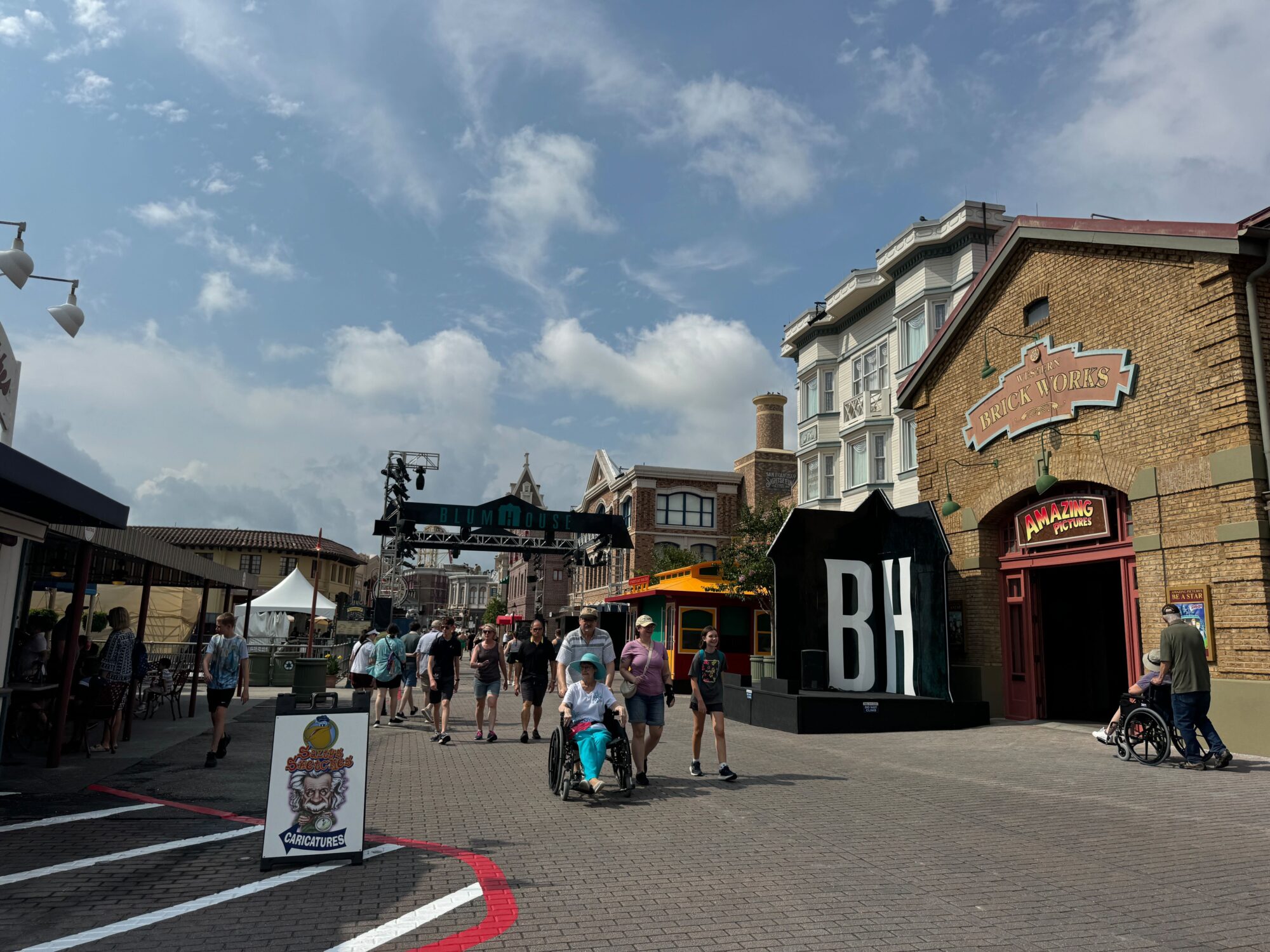 A group of people walk through a theme park area with buildings, a large "BH" structure, and a vendor booth on a sunny day with a partly cloudy sky. In the distance, the thrill of the scare zone adds an edge of excitement to their day.