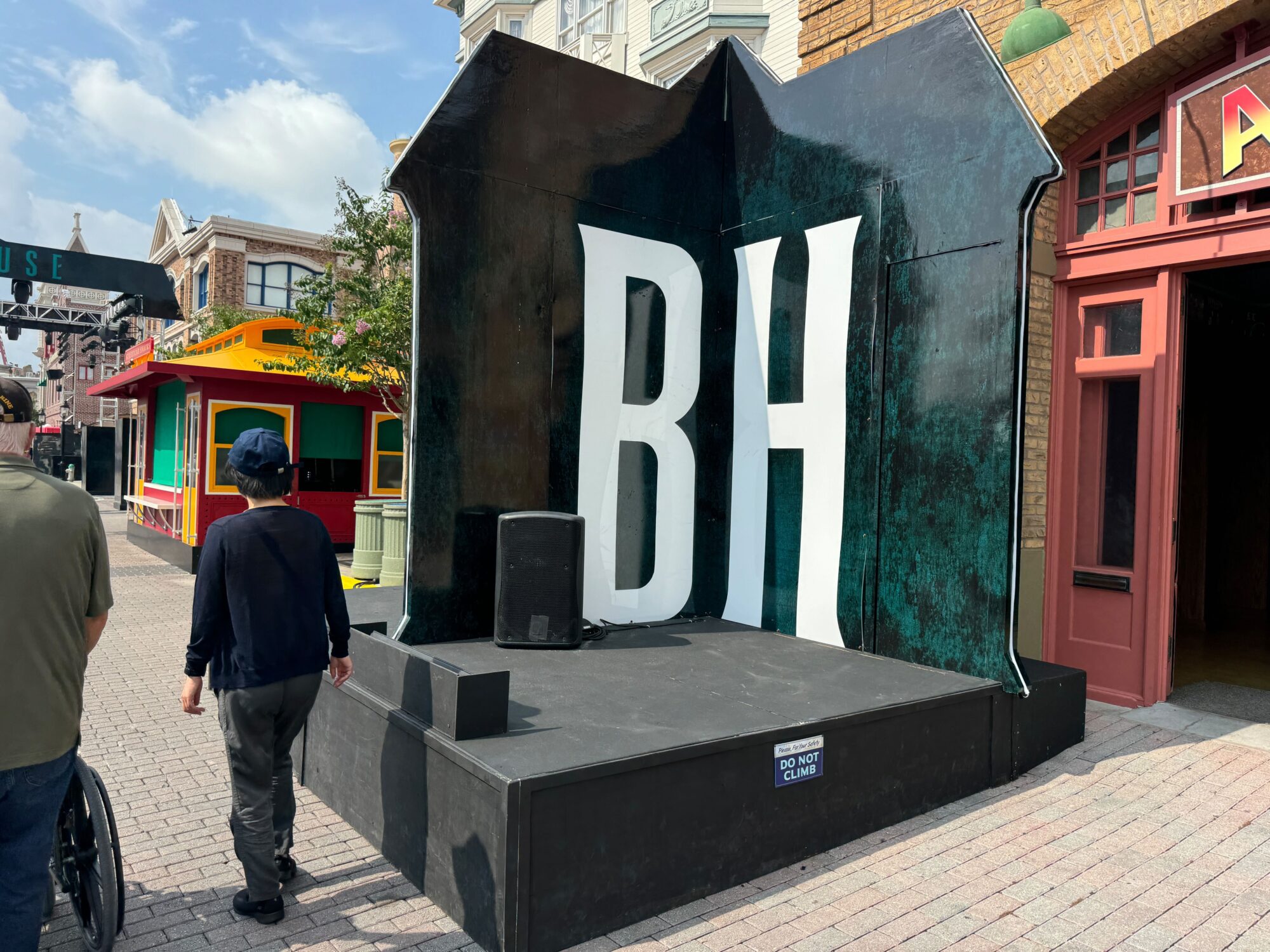 A person walks past a large black display with the letters 'BH' on it, set in an outdoor scare zone with colorful shops and buildings.