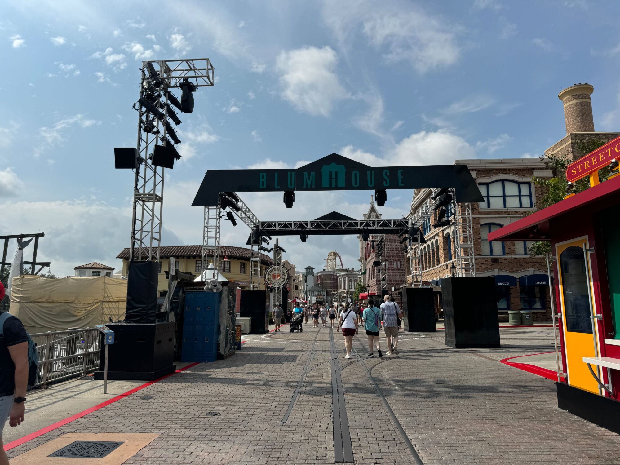 People walk along a brick-paved street under a large "Blumhouse" sign, flanked by buildings and metal trusses holding lighting equipment, transforming the area into an eerie scare zone.