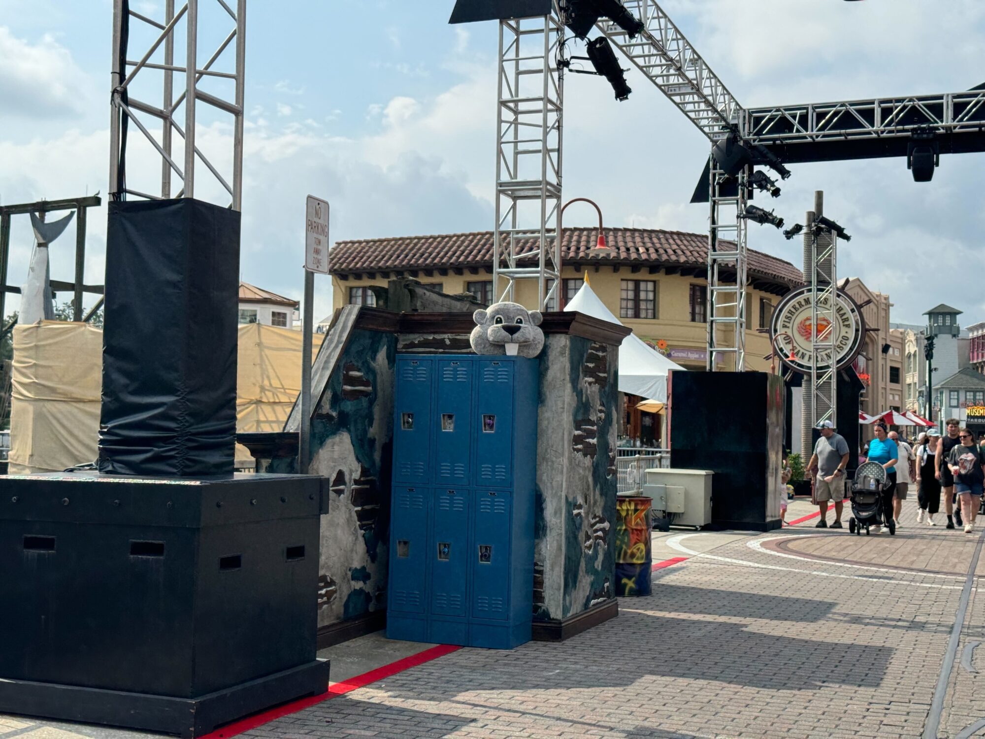 A scene with blue lockers and a carnival setup, featuring overhead trusses, and people walking in an outdoor area. A large stuffed animal sits atop the lockers, transforming the space into an unexpected scare zone.