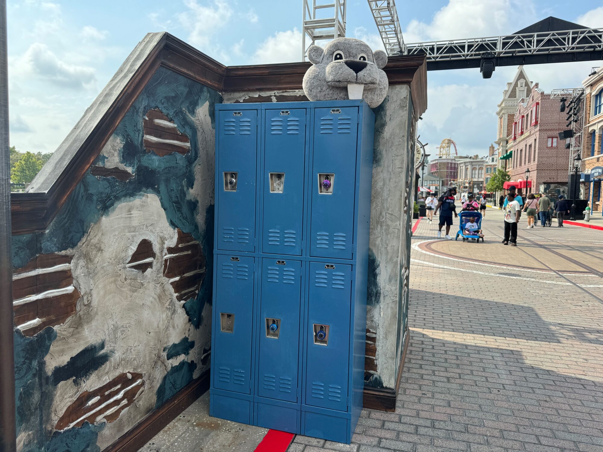 A group of blue lockers against a weathered wall with a plush walrus toy on top. In the background, people navigate a busy outdoor plaza that could easily transform into a scare zone at night.