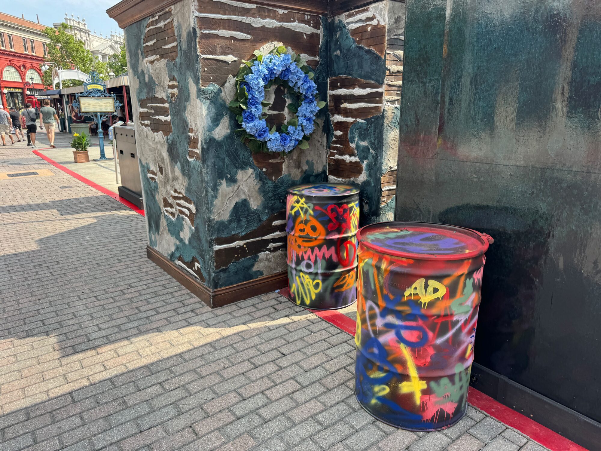 Two spray-painted barrels stand next to a weathered wall adorned with a blue flower wreath, creating a striking contrast in this urban scare zone.