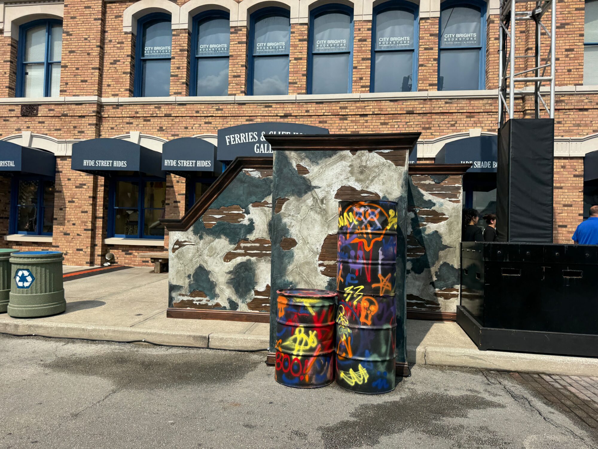 Graffiti-covered barrels are stacked beside a distressed wooden structure, with arched windows and canopy signs reading "Ferries & Gallery," "Five Street Bros.," hinting at an eerie scare zone in the background.
