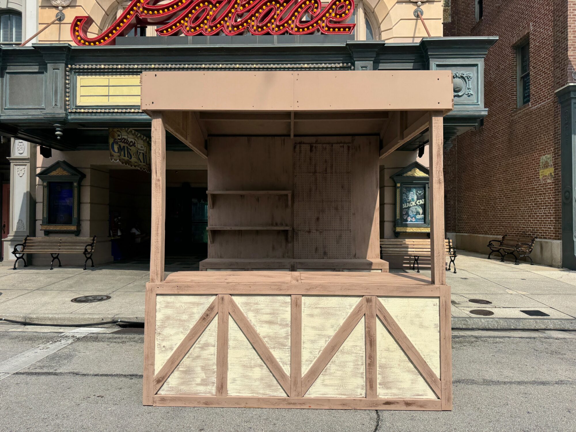 An empty wooden market stall stands on a paved street in front of a building with a "Palace" sign above, creating an eerie atmosphere reminiscent of a scare zone.