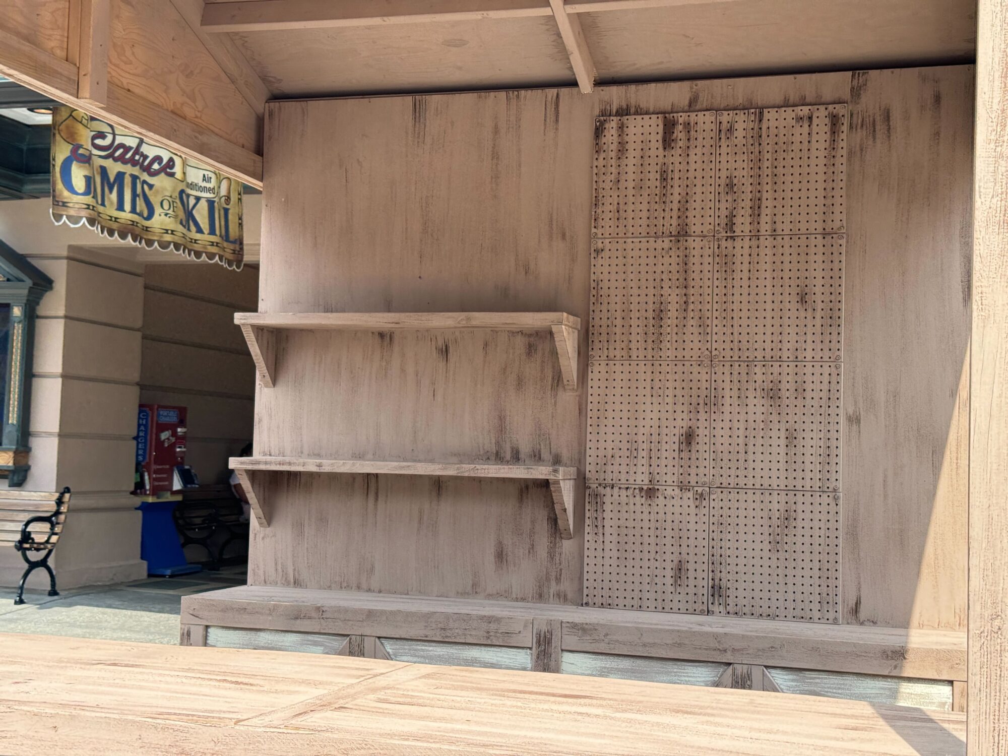 A wooden booth with empty shelves and pegboards sits beside a bench. A sign above reads "Gifts," though part of it is obscured, lending a mysterious air to this eerie scare zone.