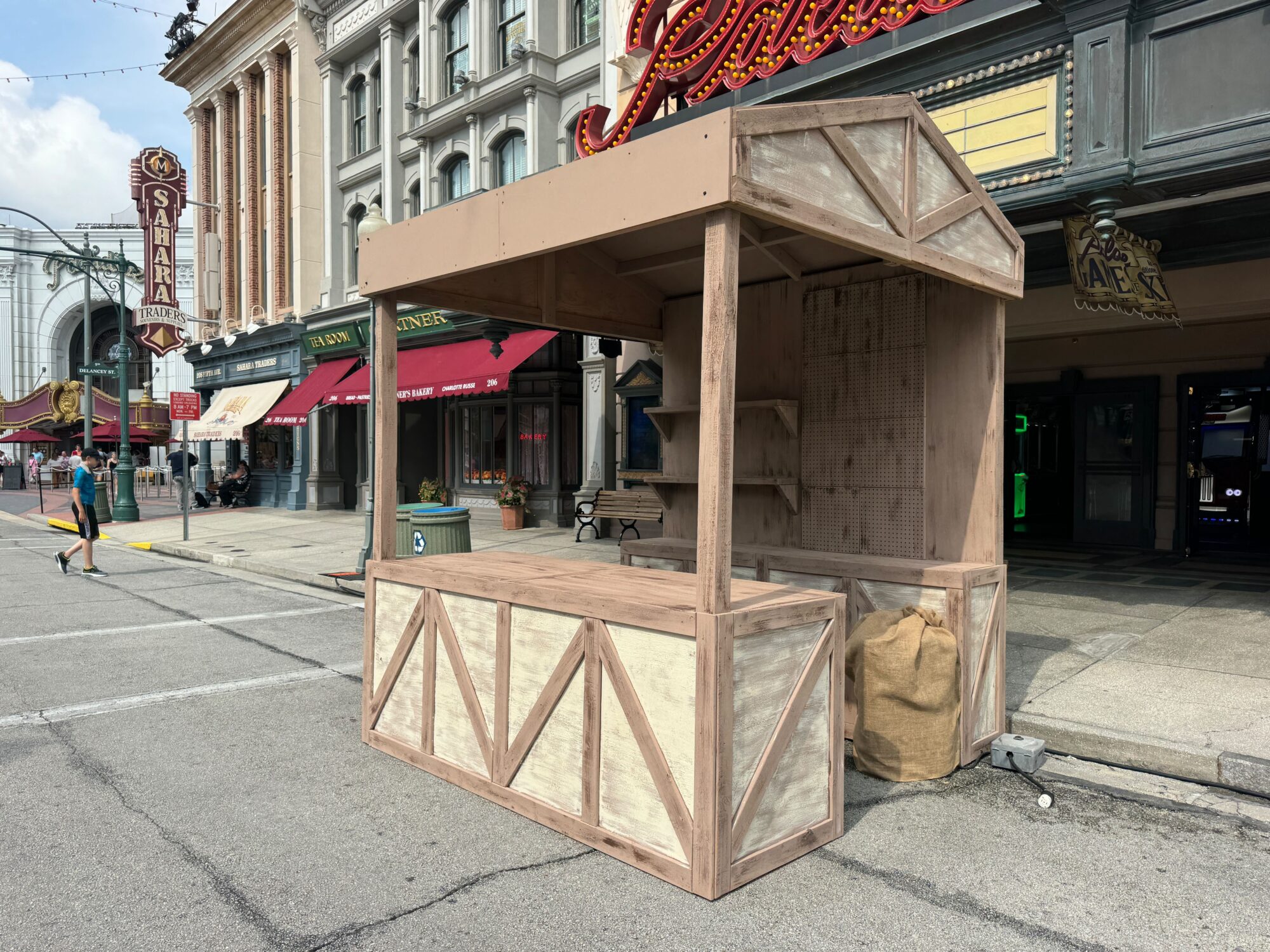 An empty wooden market stall stands on a city street, surrounded by building facades and signage, as if abandoned in a temporary scare zone.
