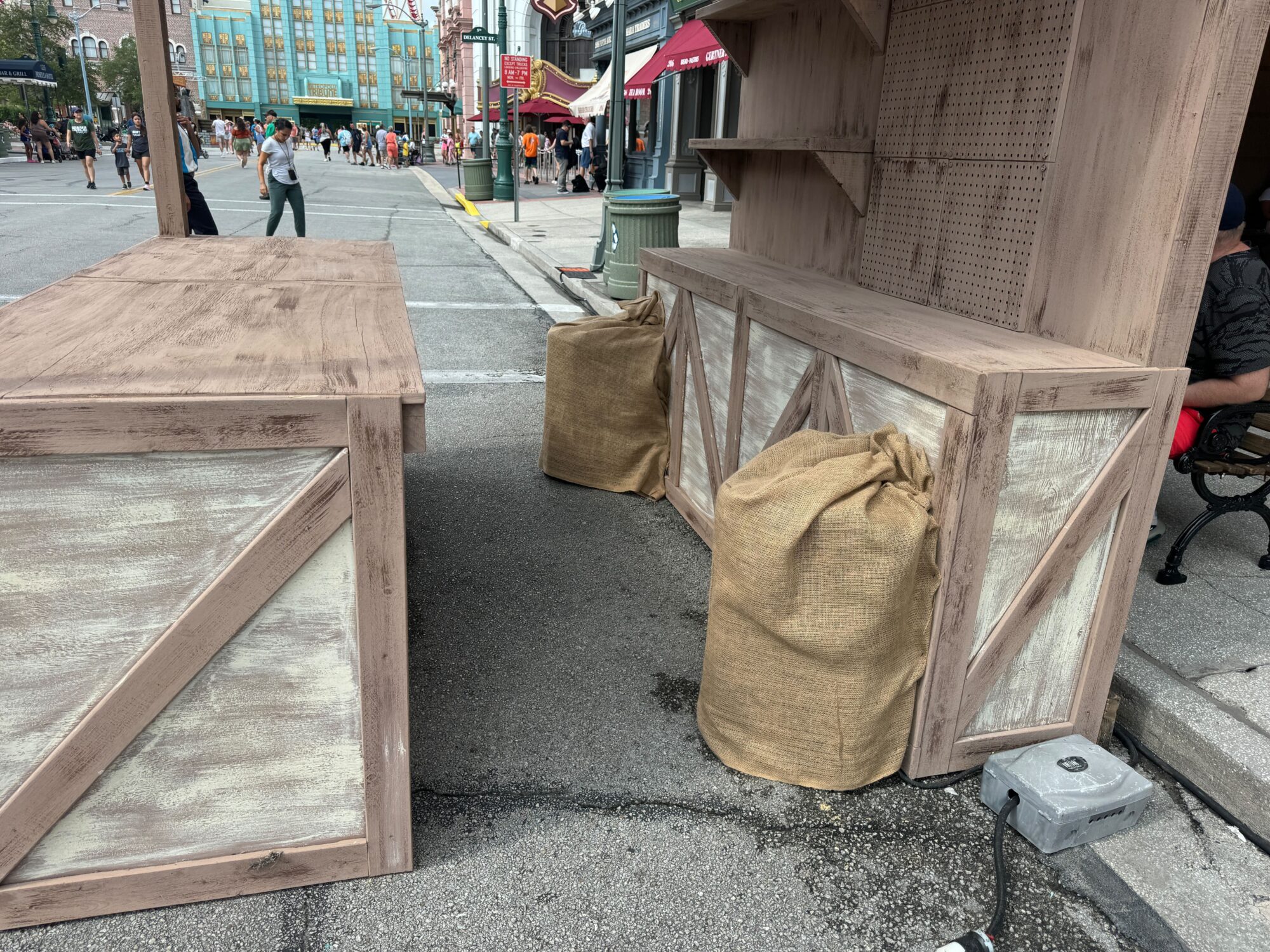 Rustic wooden tables and shelves with burlap sacks underneath are set up on a street, transforming the area into a scare zone, with people walking in the background.