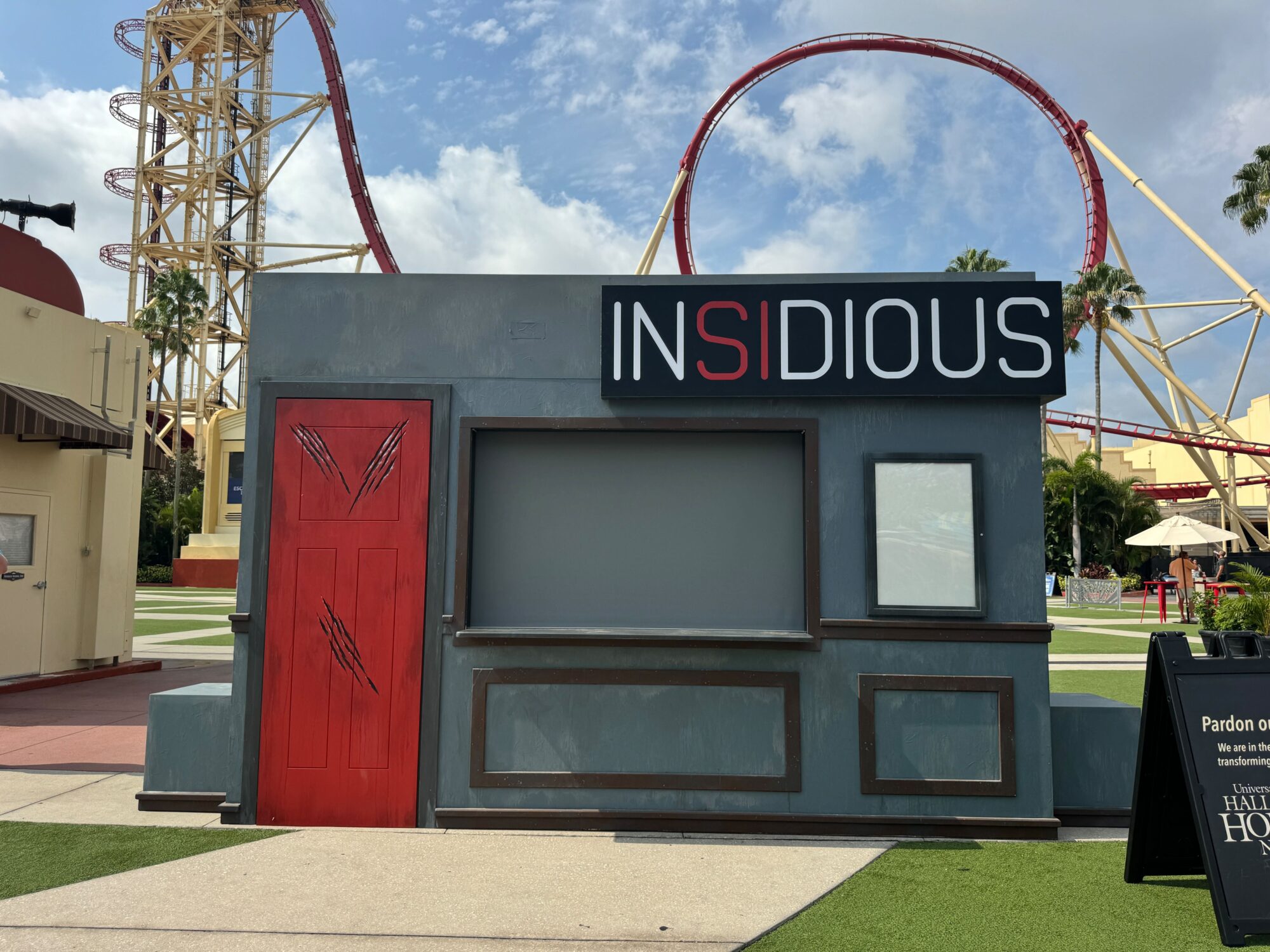 Front view of a themed photo booth labeled "Insidious" with a faux red door and scratched frames, nestled within a scare zone against an amusement park backdrop with a looping roller coaster.