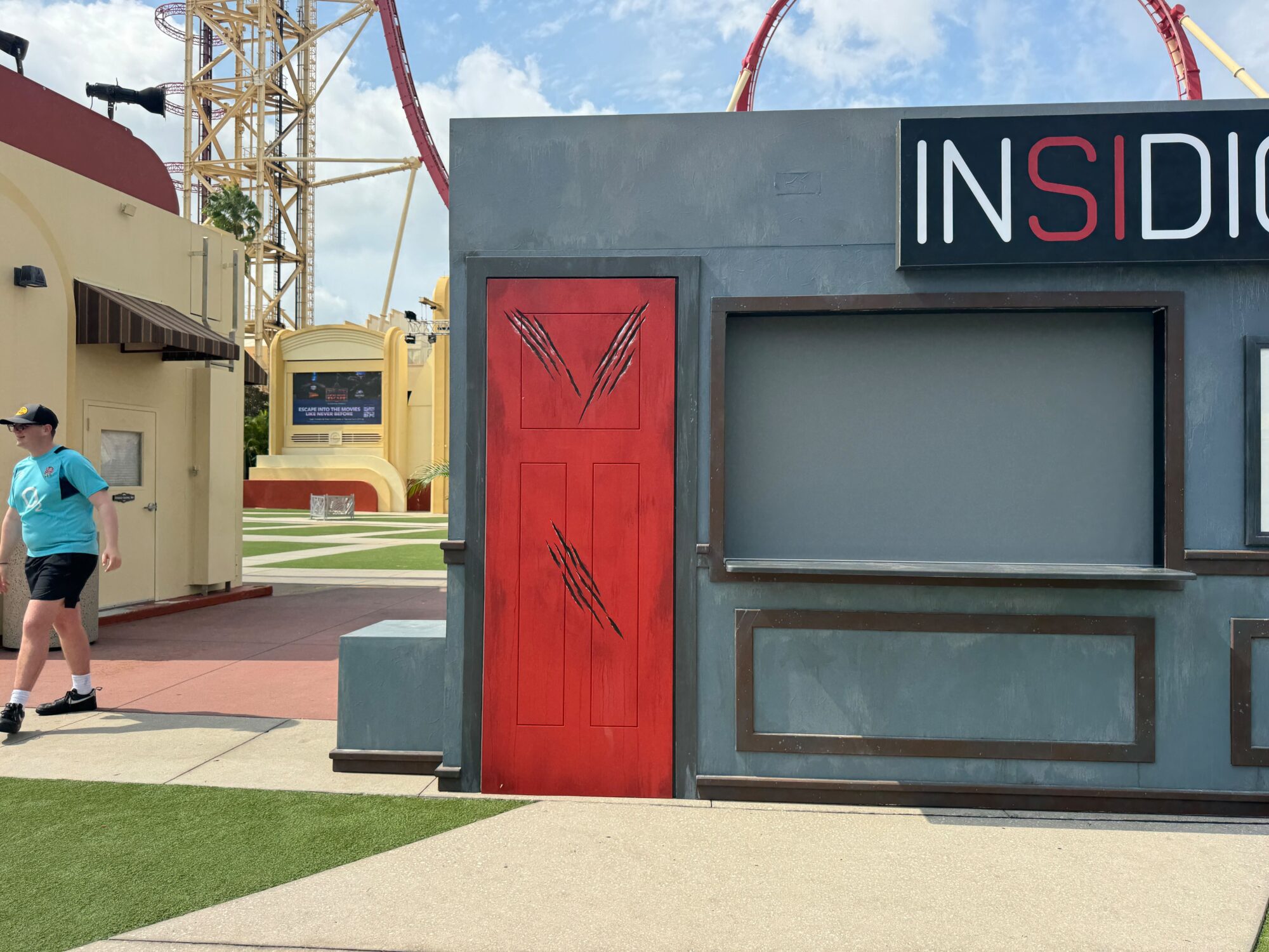 A themed attraction with a sign reading "INSIDIO" features a scratched red door and adjacent empty display window, creating an eerie scare zone. A person in a teal shirt and cap walks by on the left.