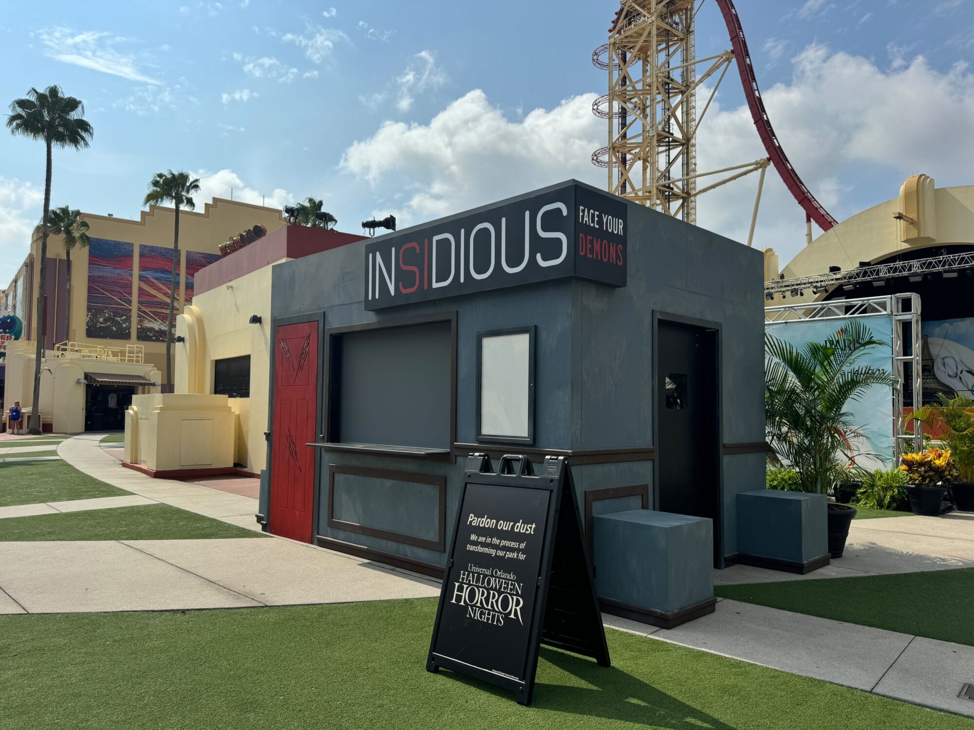 A dark, themed box office with "Insidious: Face Your Demons" signage and a construction sign for Halloween Horror Nights stands in a theme park scare zone, with palm trees and a roller coaster in the background.