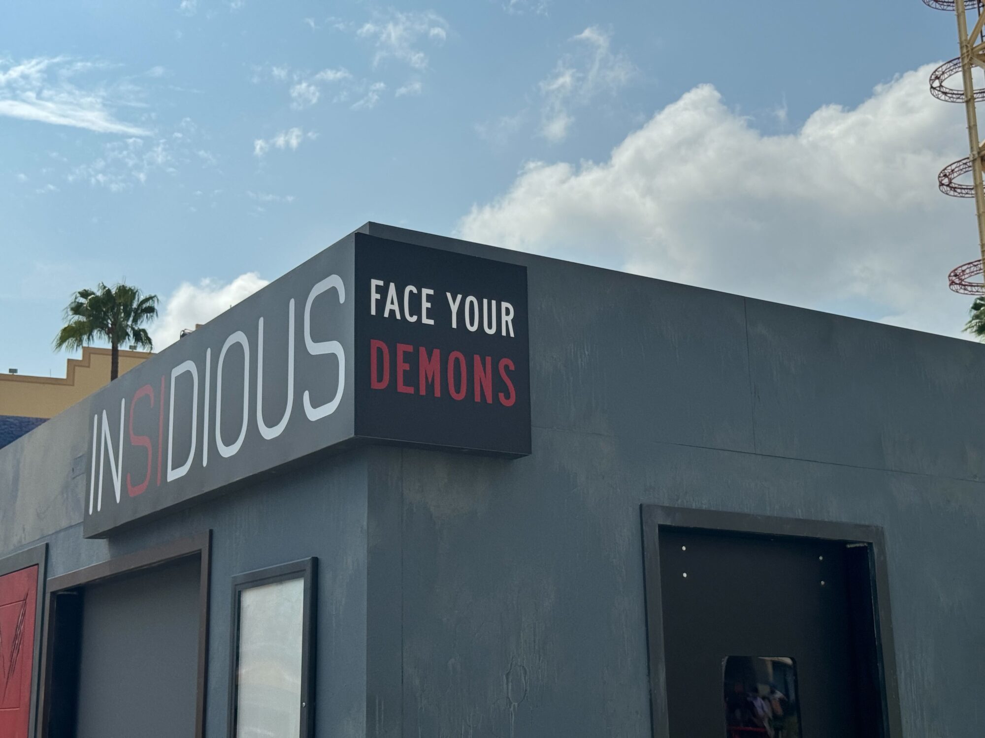 A building with a sign reading "Insidious" and "Face Your Demons" on its facade, set against a partly cloudy sky with a glimpse of a roller coaster and palm trees in the background, marks the entrance to an eerie scare zone.