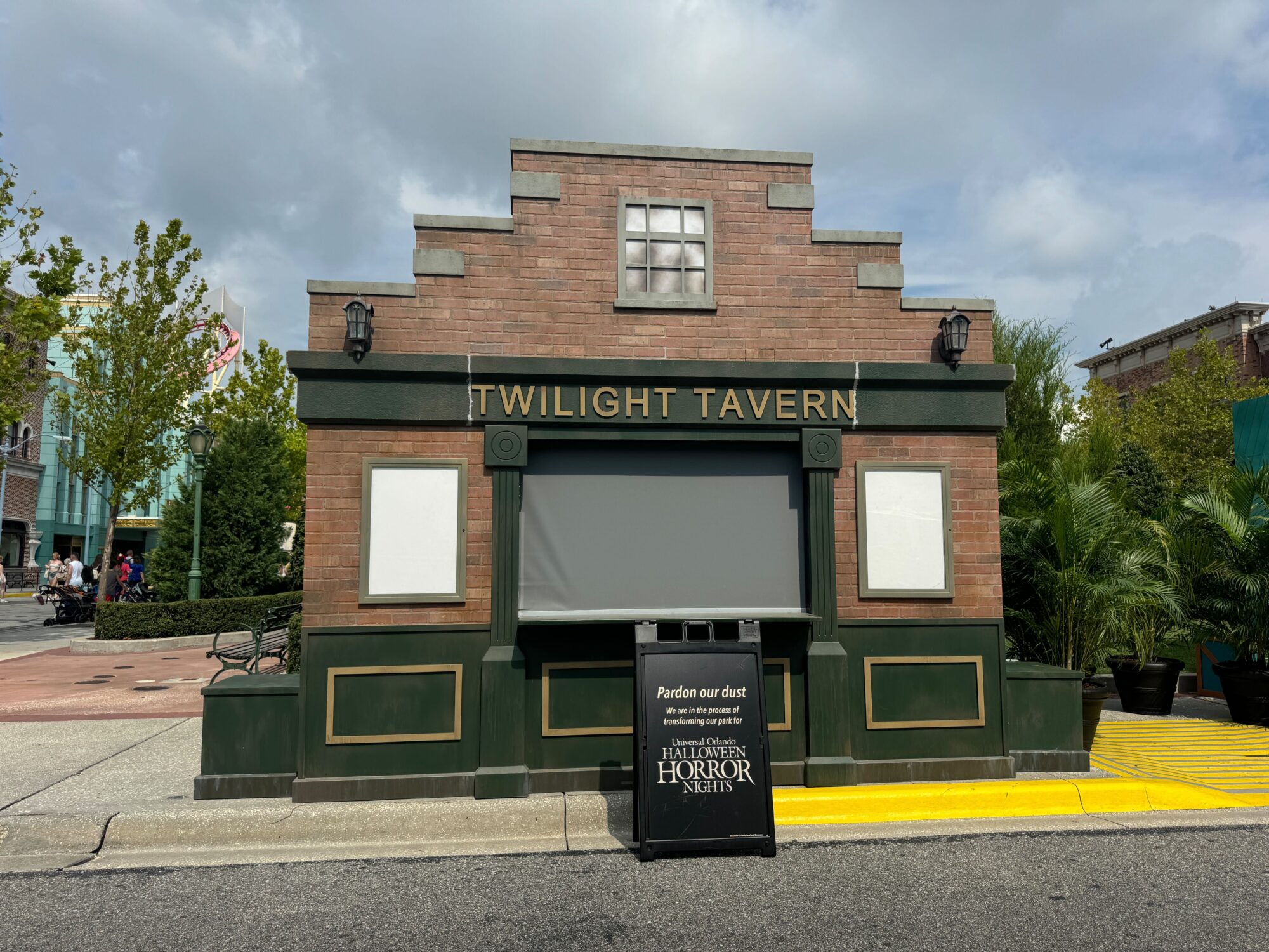 A brick building with a sign reading "Twilight Tavern" is closed, with a construction sign in front stating "Pardon our dust." The scene, reminiscent of a spooky scare zone, is outdoors with trees and a cloudy sky in the background.