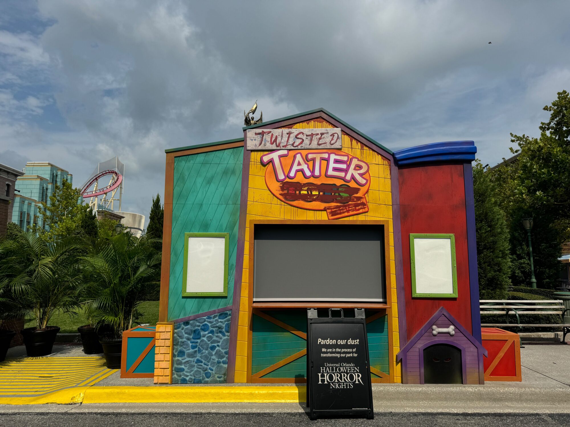 A colorful stand named "Twisted Tater," under construction with a "Pardon our dust" sign in front, sits against a cloudy sky and nearby greenery in the heart of the new scare zone.