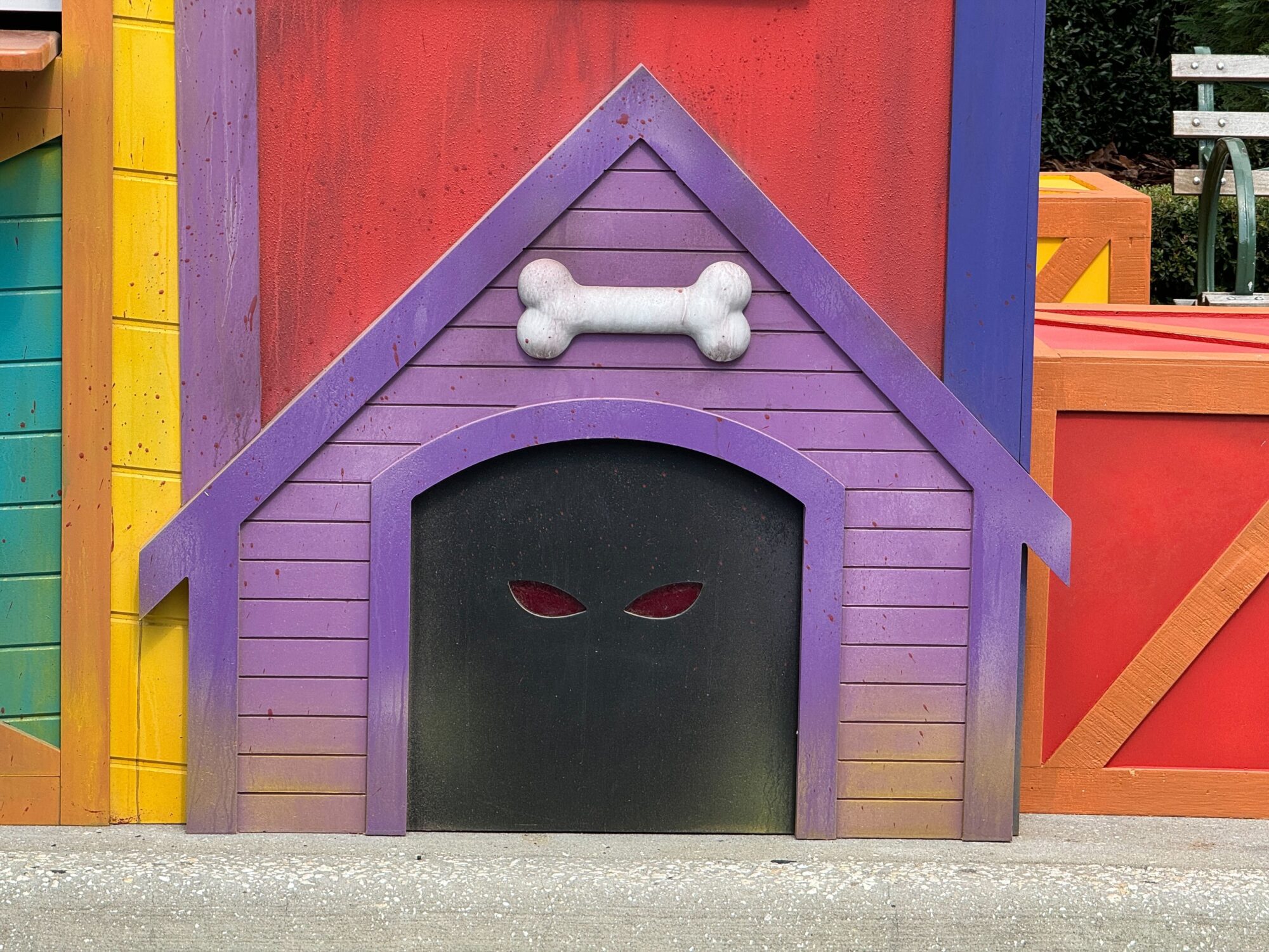 A colorful dog house with a purple facade, featuring a white bone decoration above the entrance. The entrance is painted black with red eyes, creating a spooky effect, perfect for transforming any yard into a mini scare zone.