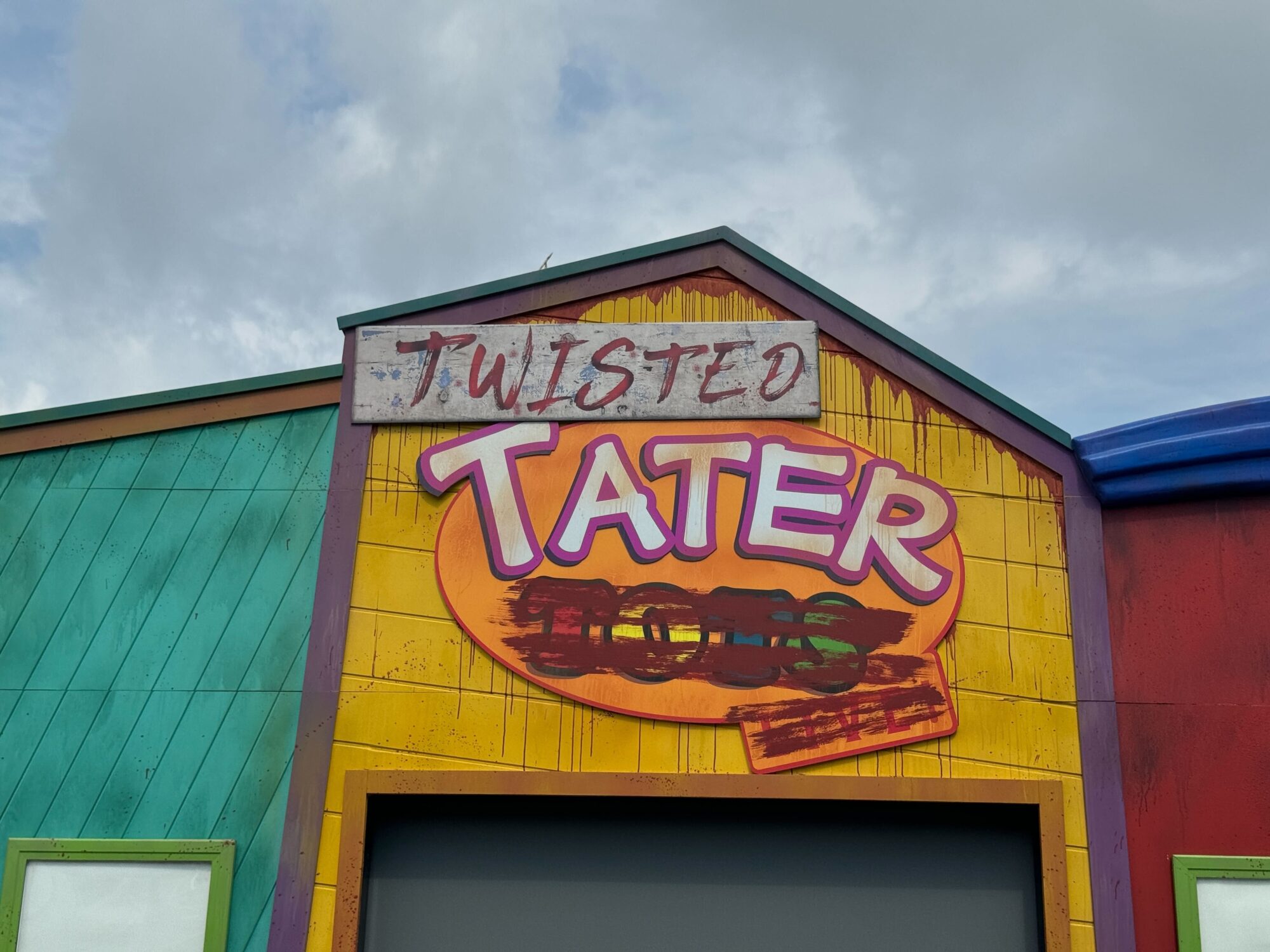 Colorful building facade with a sign that reads "Twisted Tater," featuring a large, stylized potato with a skewer through it. The background reveals a cloudy sky, hinting at the nearby scare zone and adding an eerie touch to the scene.