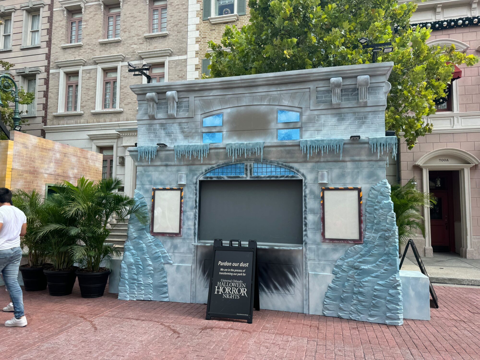 Facade of a themed attraction with an icy exterior, empty frames, and a sign reading "Pardon our dust" and "Halloween Horror Nights." A person is partly visible on the left, setting the eerie tone for this scare zone.