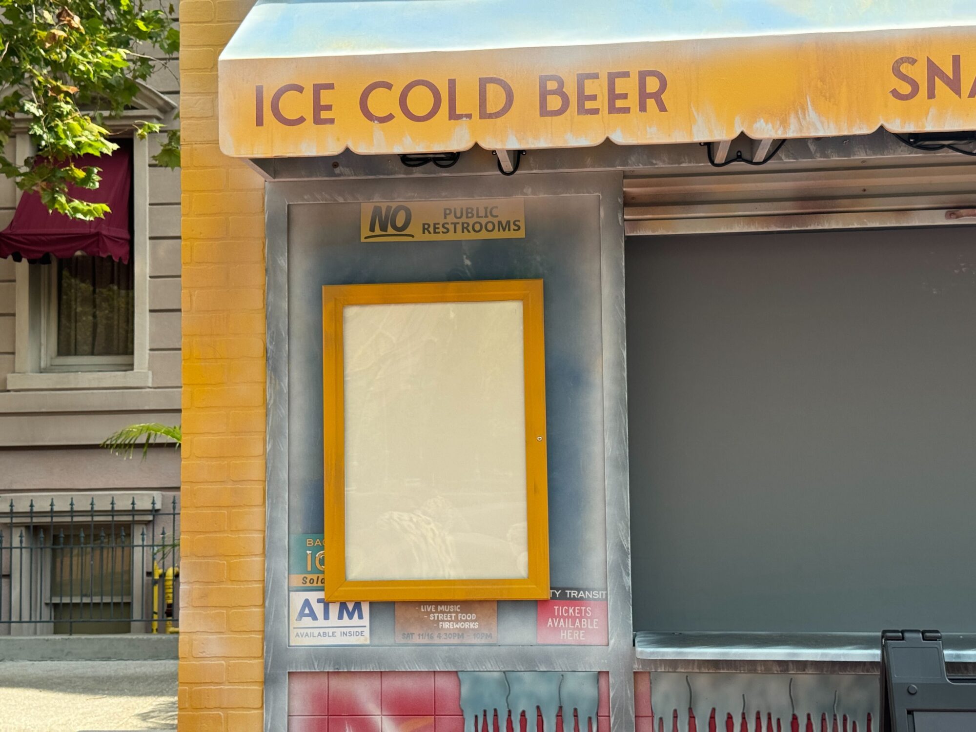 Food stand with a yellow-framed empty display under a sign saying "ICE COLD BEER." Positioned within the scare zone, another sign indicates "NO PUBLIC RESTROOMS." Other signage mentions tickets, ATM, and various refreshments.