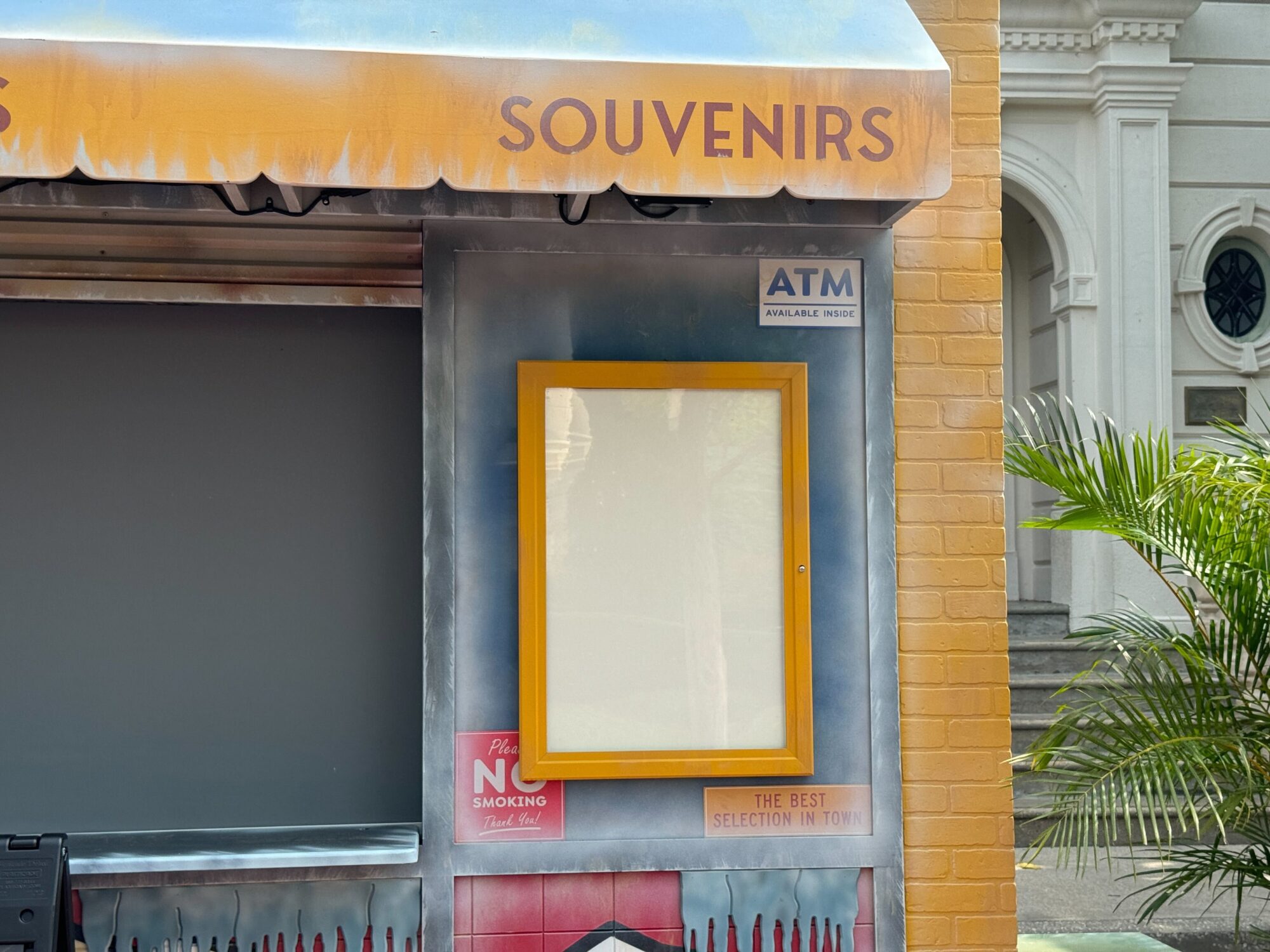 A closed souvenir shop with an ATM sign, no smoking sign, and empty display sits eerily in a scare zone. The awning reads "SOUVENIRS," set against a yellow wall with a nearby plant.