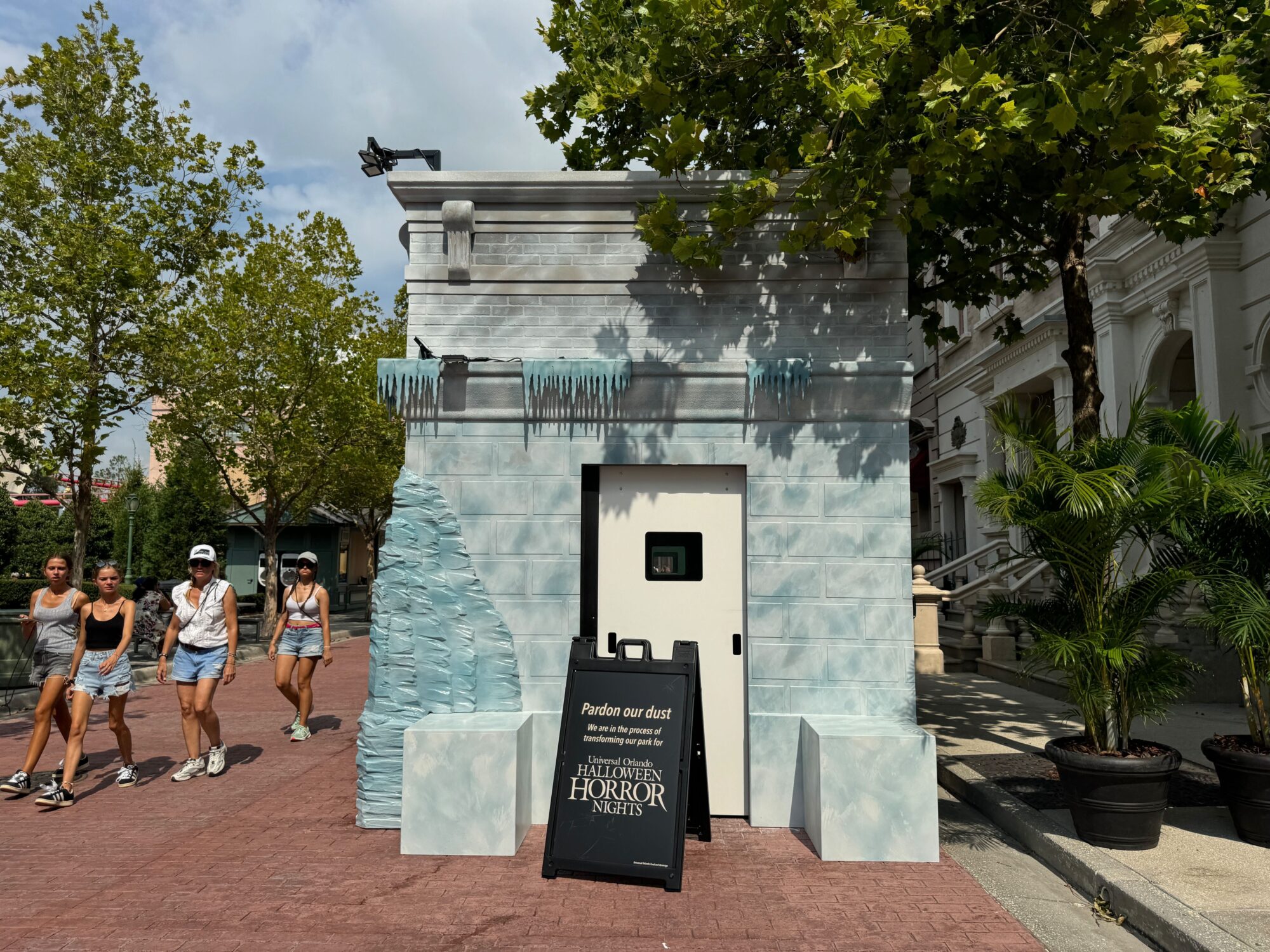 A small, ice-themed construction booth with a sign reading "Pardon our dust" for the Halloween Horror Nights scare zone stands on a brick pathway. Several people walk by under a tree-covered sky.
