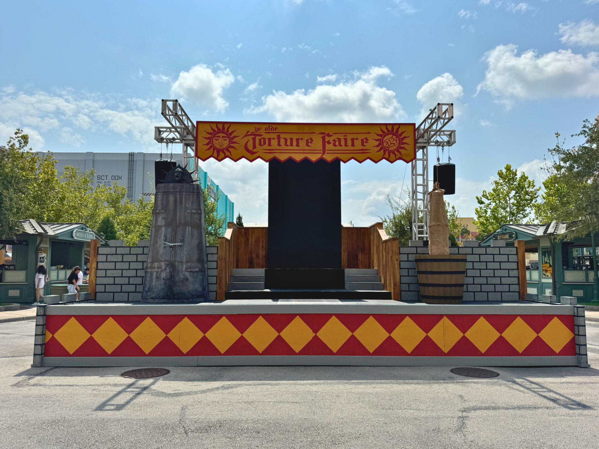 An outdoor stage setup for “The Torture Faire,” featuring a red and gold banner, platform with a black backdrop, two side towers, and decorative brickwork. Trees and booths are visible in the background, creating an eerie scare zone atmosphere.