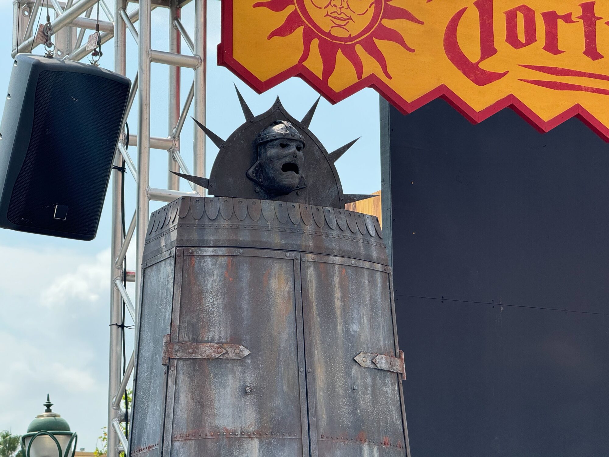 A stylized statue with a helmeted head and spiked crown emerges from a large metallic cylinder, part of a themed scare zone under a sign with ornate script and sun icon.