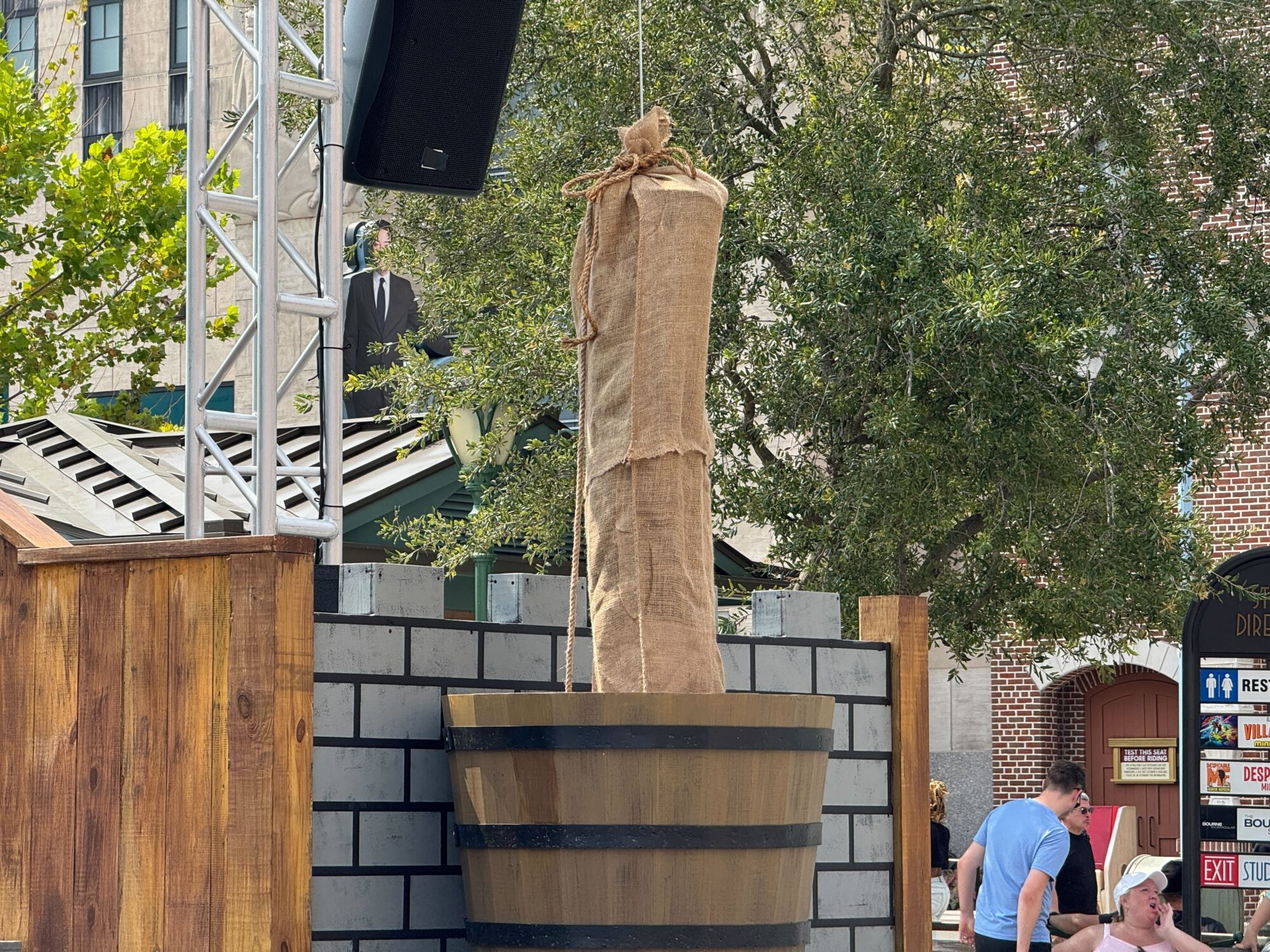 A burlap-covered object is suspended above a large wooden barrel set within a constructed brick structure, creating an eerie scare zone. Trees and people, including one in a pink shirt, are visible in the background.