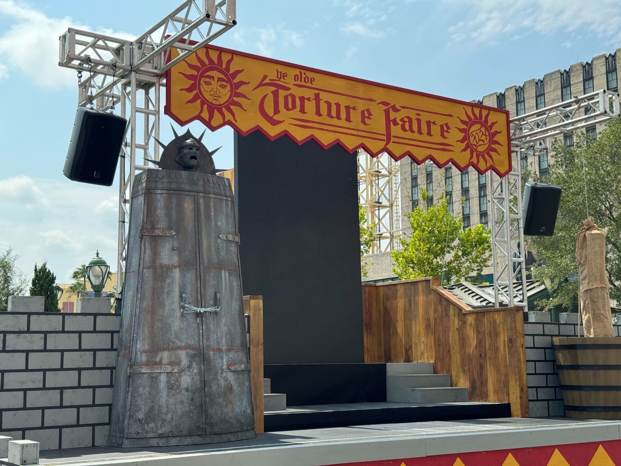 A stage setup with a banner reading "Ye Olde Torture Faire" transforms the area into a chilling scare zone. Props include a metal cage and wooden structure, all set against a backdrop of buildings and trees under a partly cloudy sky.
