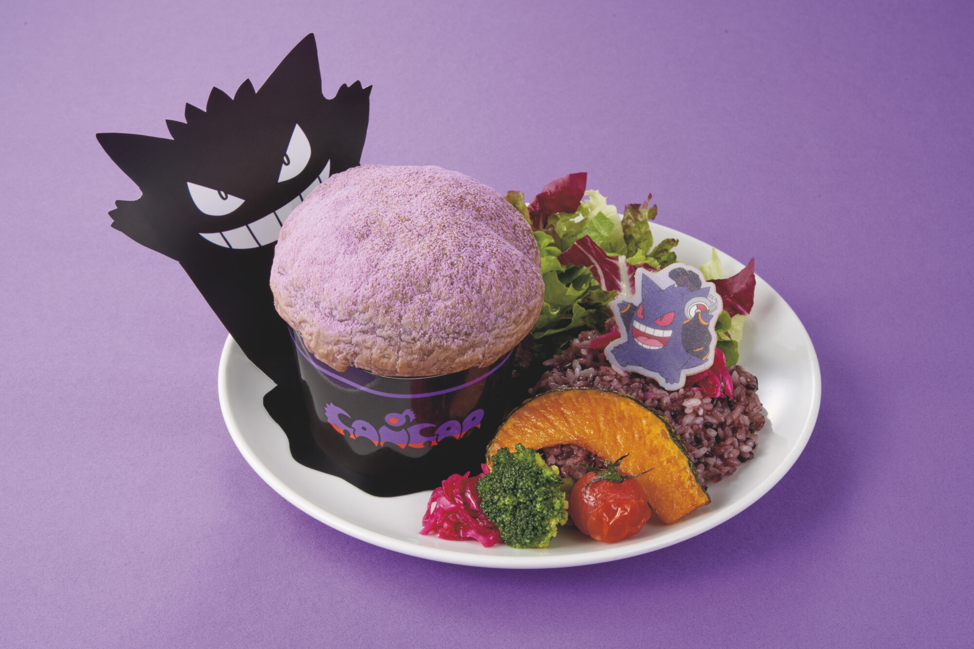 A purple burger on a white plate with a side of mixed greens, shaped rice, and vegetables. The food is themed with a black and purple cartoon character. Background is purple.