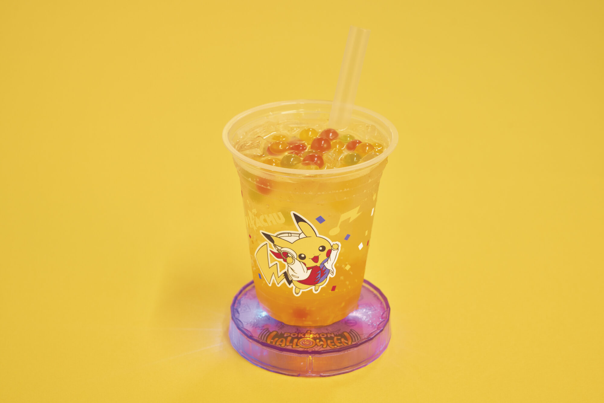A plastic cup featuring Pikachu filled with a yellow drink and boba pearls, topped with a lid and two straws, placed on a purple coaster against a yellow background.