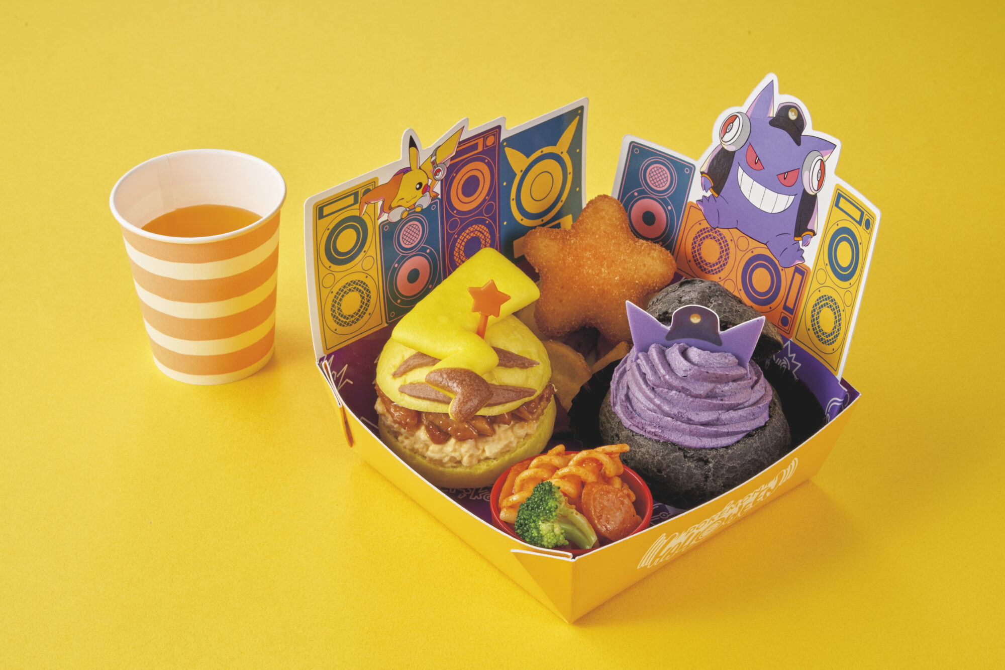 A themed lunch box with various colorful foods, including a burger with a yellow star decoration, purple mashed potatoes, a star-shaped item, and vegetables, beside a striped cup of tea, on a yellow background.
