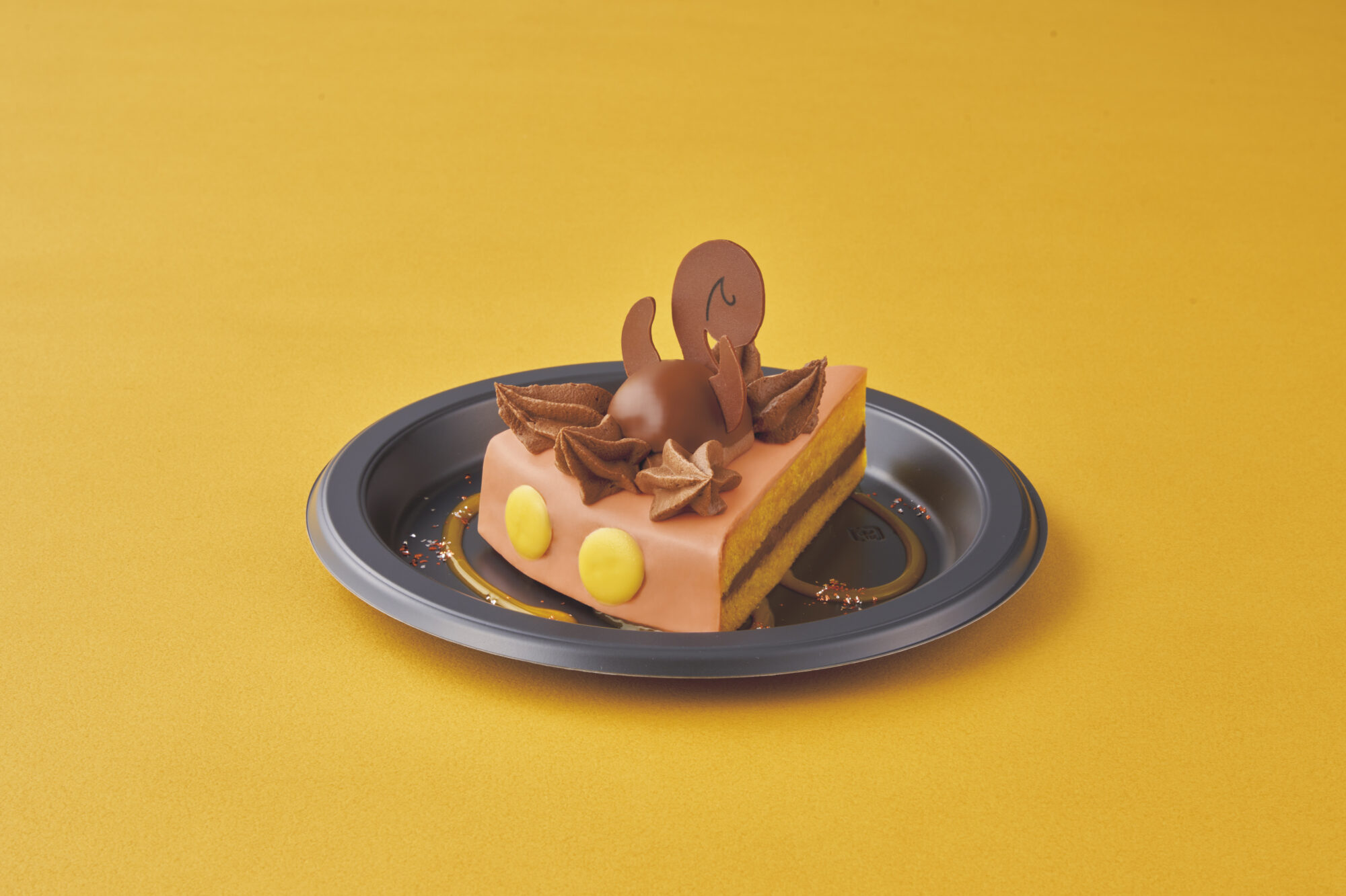 A rectangular piece of cake with yellow frosting on the sides, topped with brown chocolate decorations and a small smiling chocolate figure, served on a black plate against a yellow background.