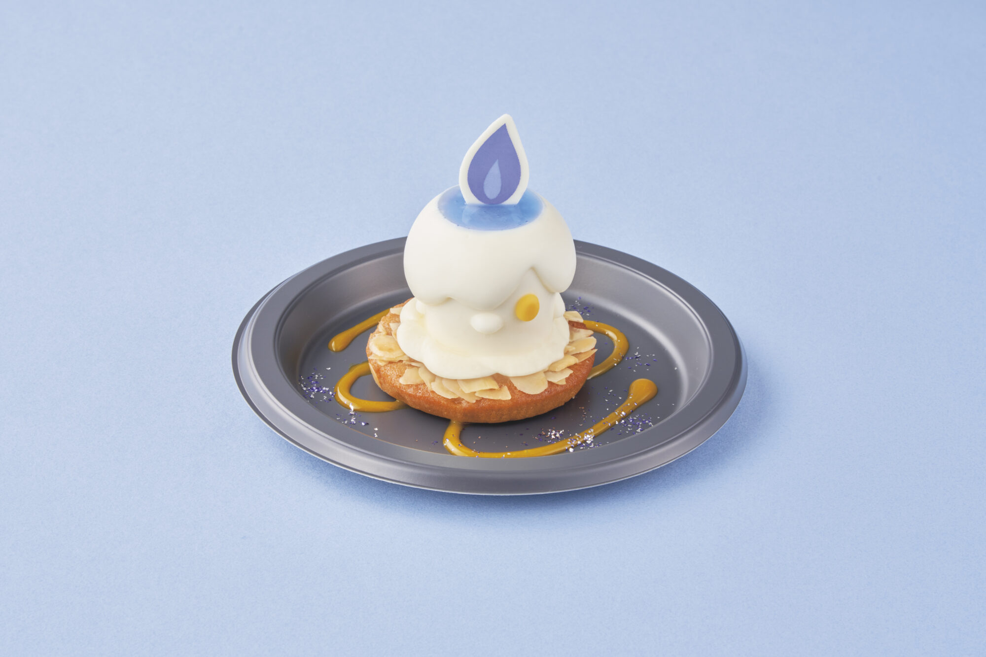 A dessert resembling a candle with white cream on top of a pastry, almond slices, and a blue flame decoration, placed on a black plate with a swirl of yellow sauce on a light blue background.