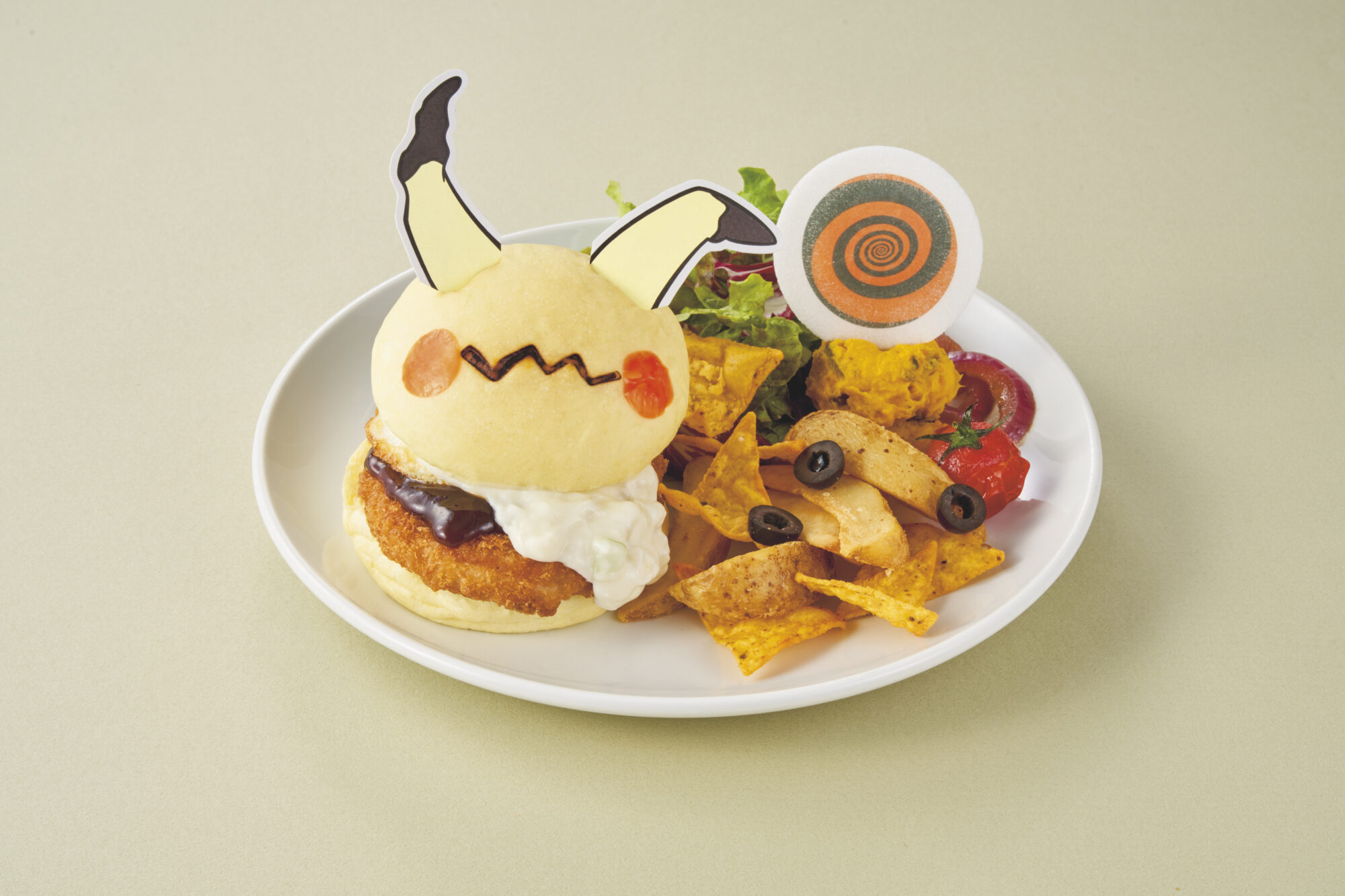 A themed burger resembling a character, garnished with sauce and a dollop of cream, served with nachos, potato wedges, a small salad, and a round cookie featuring a swirl pattern.