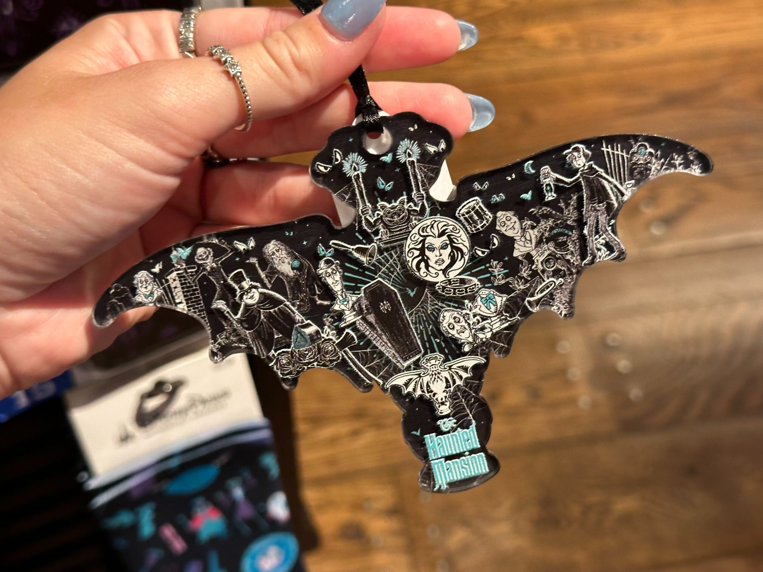 A hand holding a bat-shaped Haunted Mansion ornament with intricate designs and characters from the attraction against a wooden background, showcasing perfect Halloween merchandise.