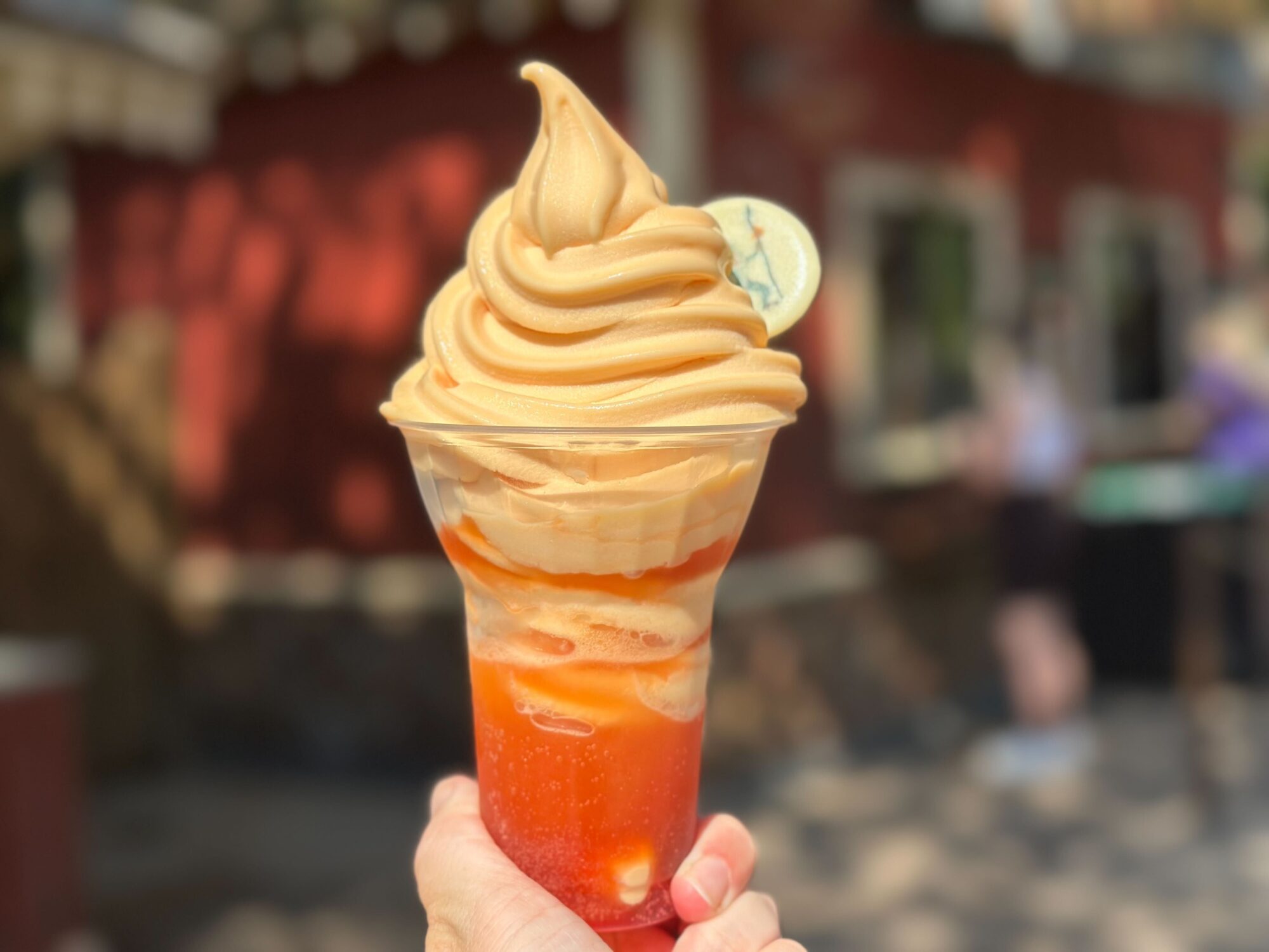 A hand holds a plastic cup filled with swirled soft serve ice cream on top of a red-orange beverage, with a small slice of lime on the rim. The background is blurred with outdoor scenery.