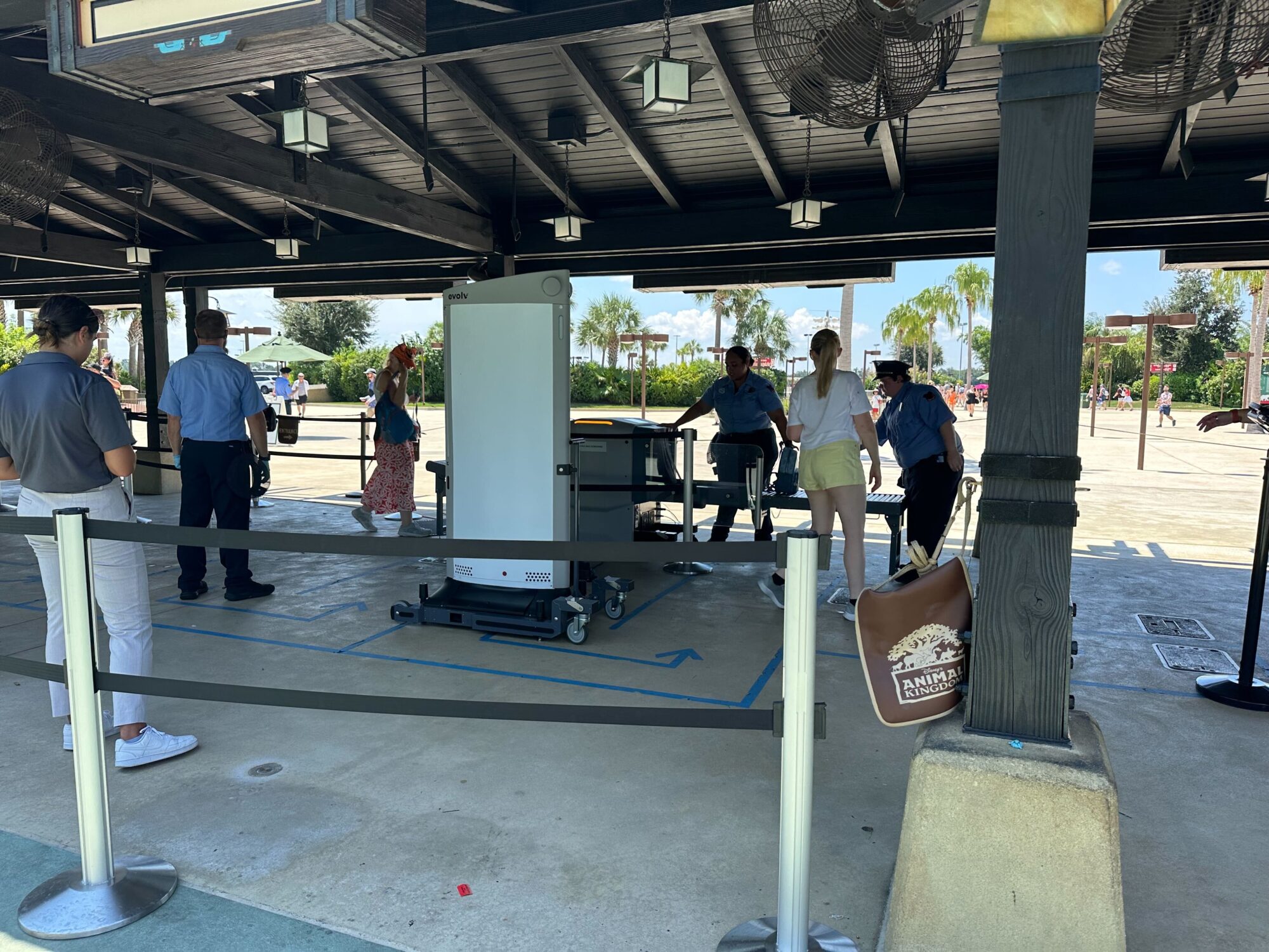 Security checkpoint with several guests waiting to go through x-ray machines under a shaded structure. Two security personnel are assisting. Nearby, a person holds a drink and a bag labeled "Animal," adding to the lively atmosphere reminiscent of Disney World.