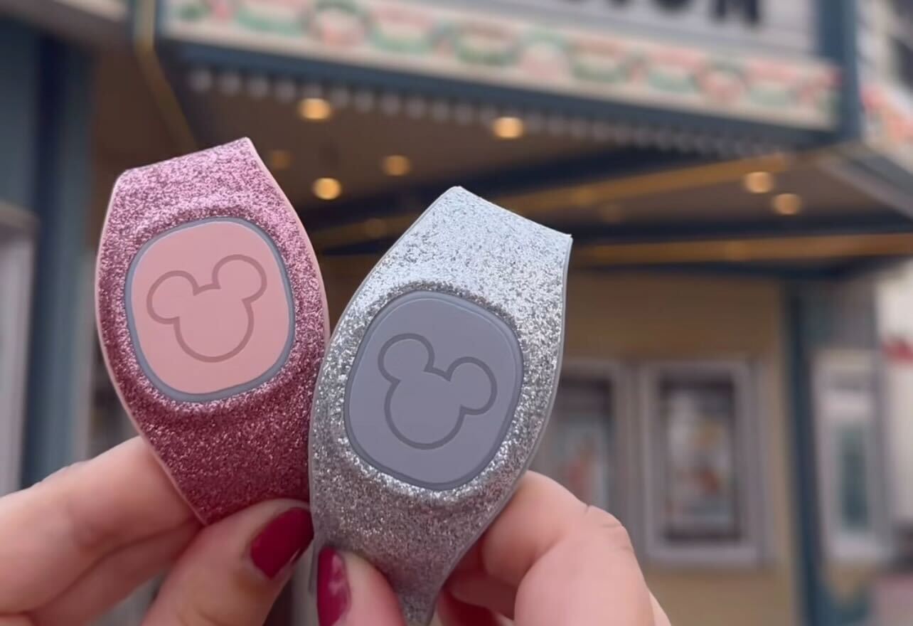 Two hands hold new MagicBands with Mickey Mouse logos, one pink and one silver, in front of a lit marquee.