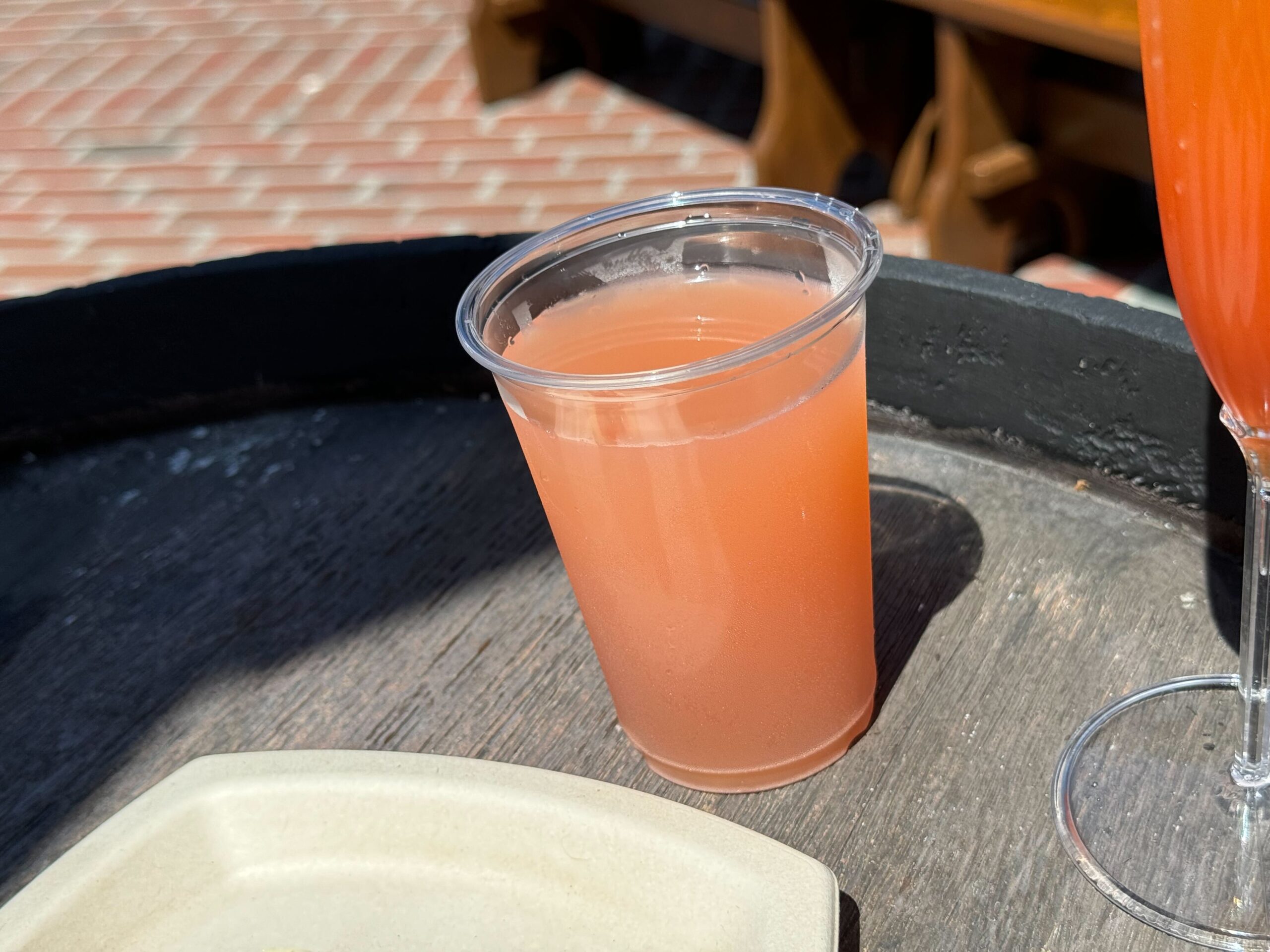 A clear plastic cup filled with a pink beverage sits on a wooden surface next to a glass with a similar drink and a beige plate, evoking an elegant scene straight out of Food & Wine.