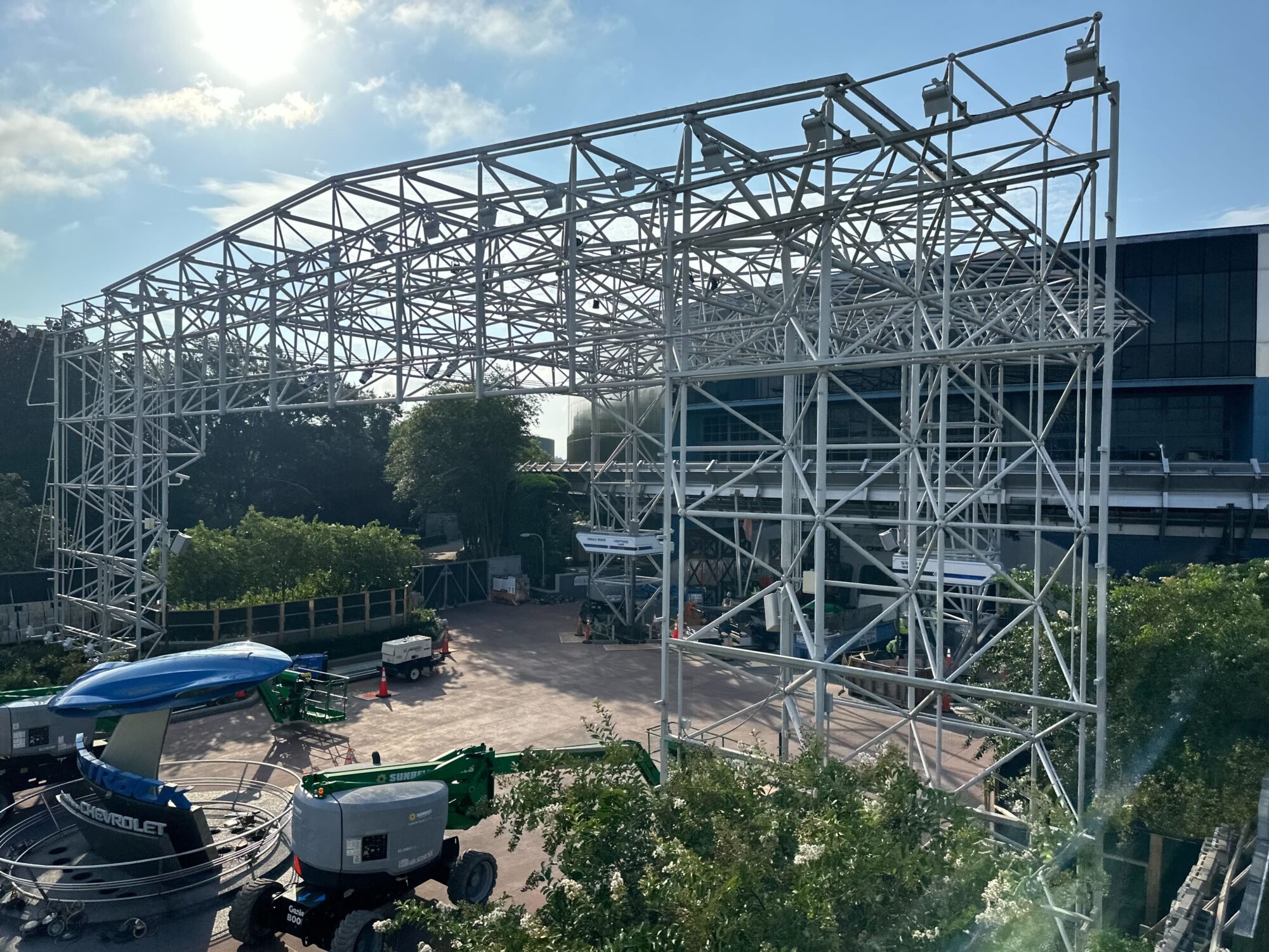 A large metal framework structure is under construction outdoors, with smaller safety equipment and machinery around it. Trees and a building are visible in the background under a partly cloudy sky.