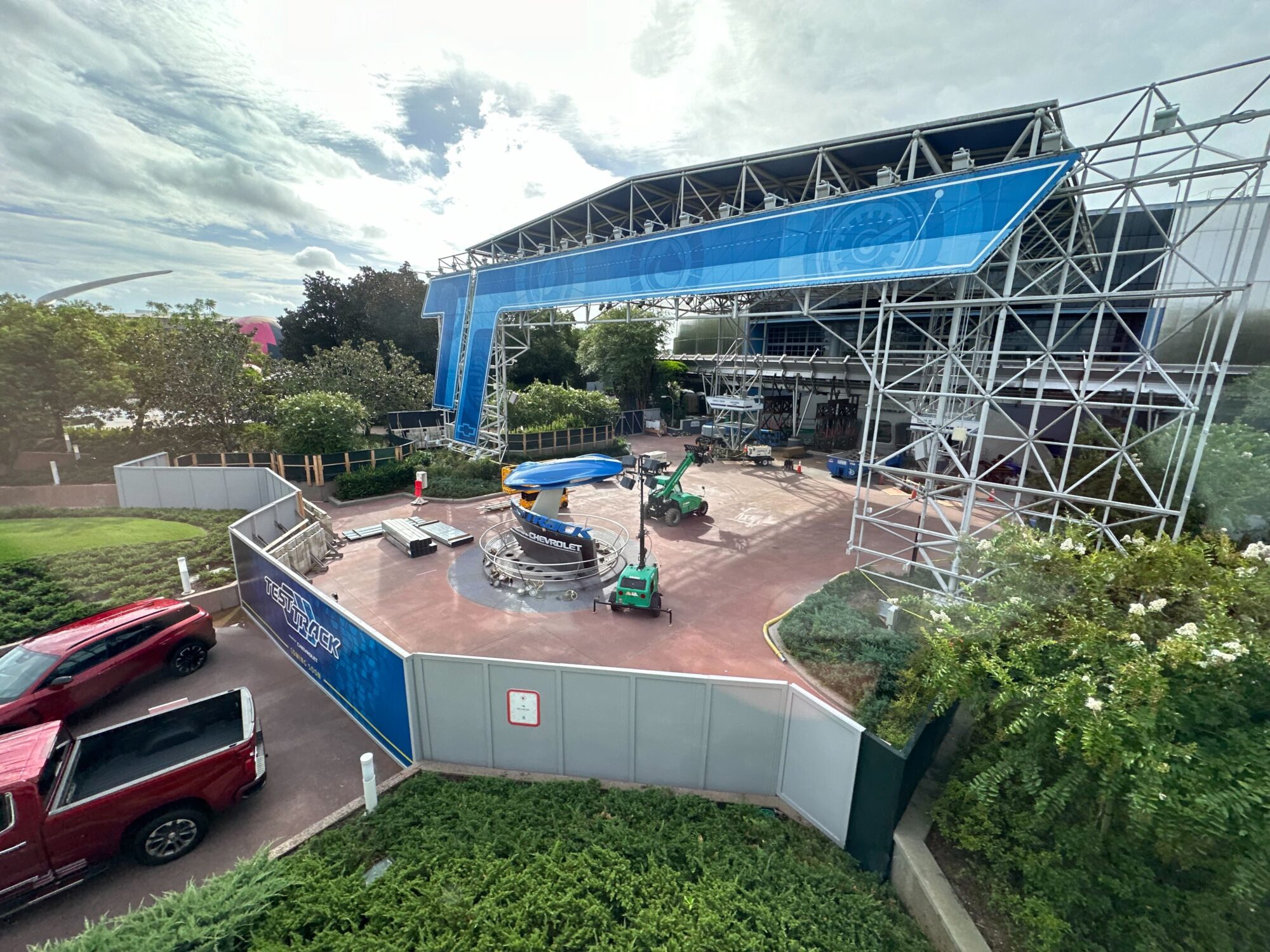 A construction site with scaffolding, vehicles, and machinery set up around an unfinished structure. A large blue and white banner is displayed above the site, and greenery surrounds the area.