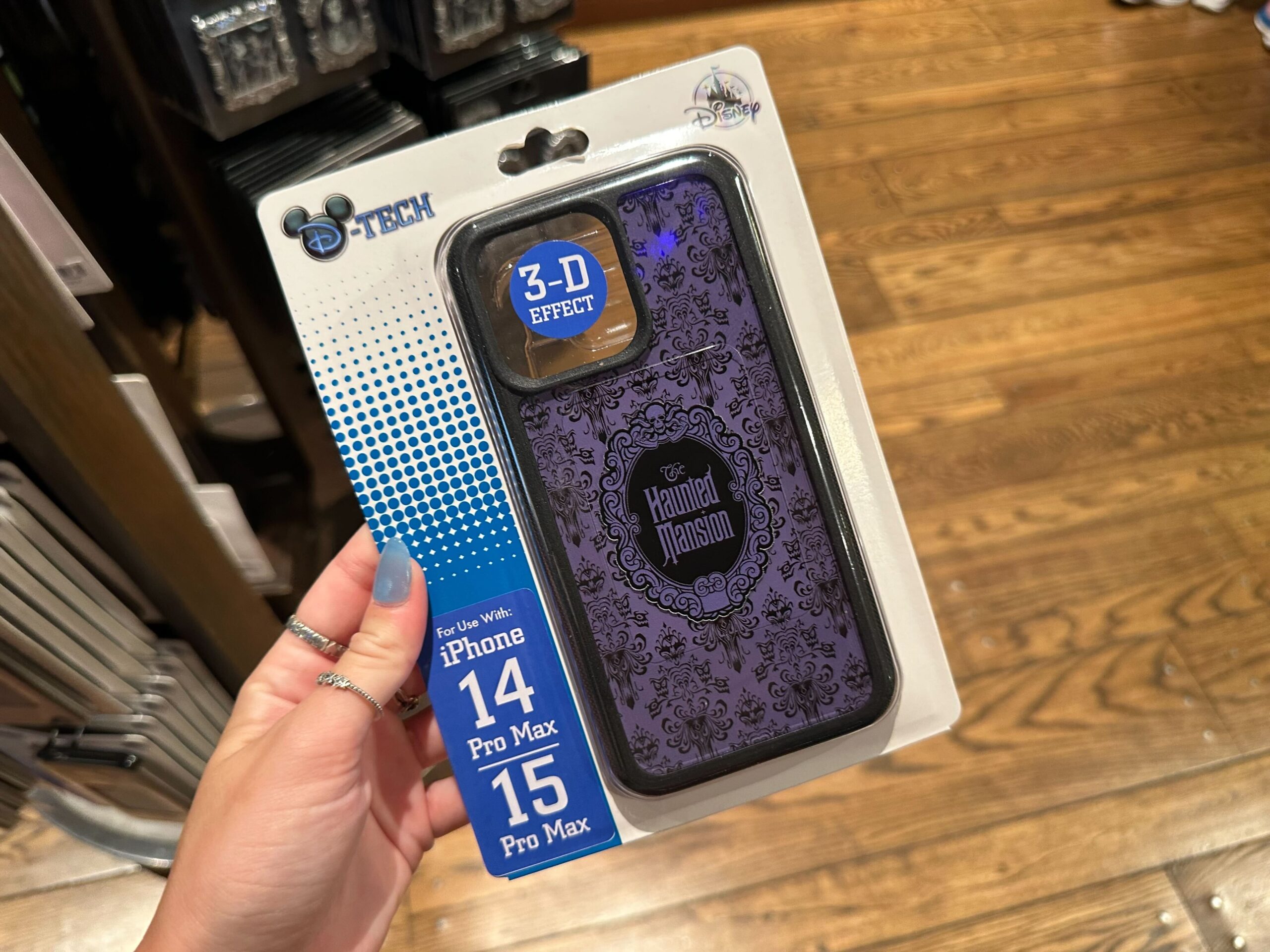 A hand holds a packaged 3-D effect iPhone case compatible with iPhone 14 Pro Max and 15 Pro Max. This piece of Halloween merchandise features "The Haunted Mansion" theme in eerie black and purple colors.
