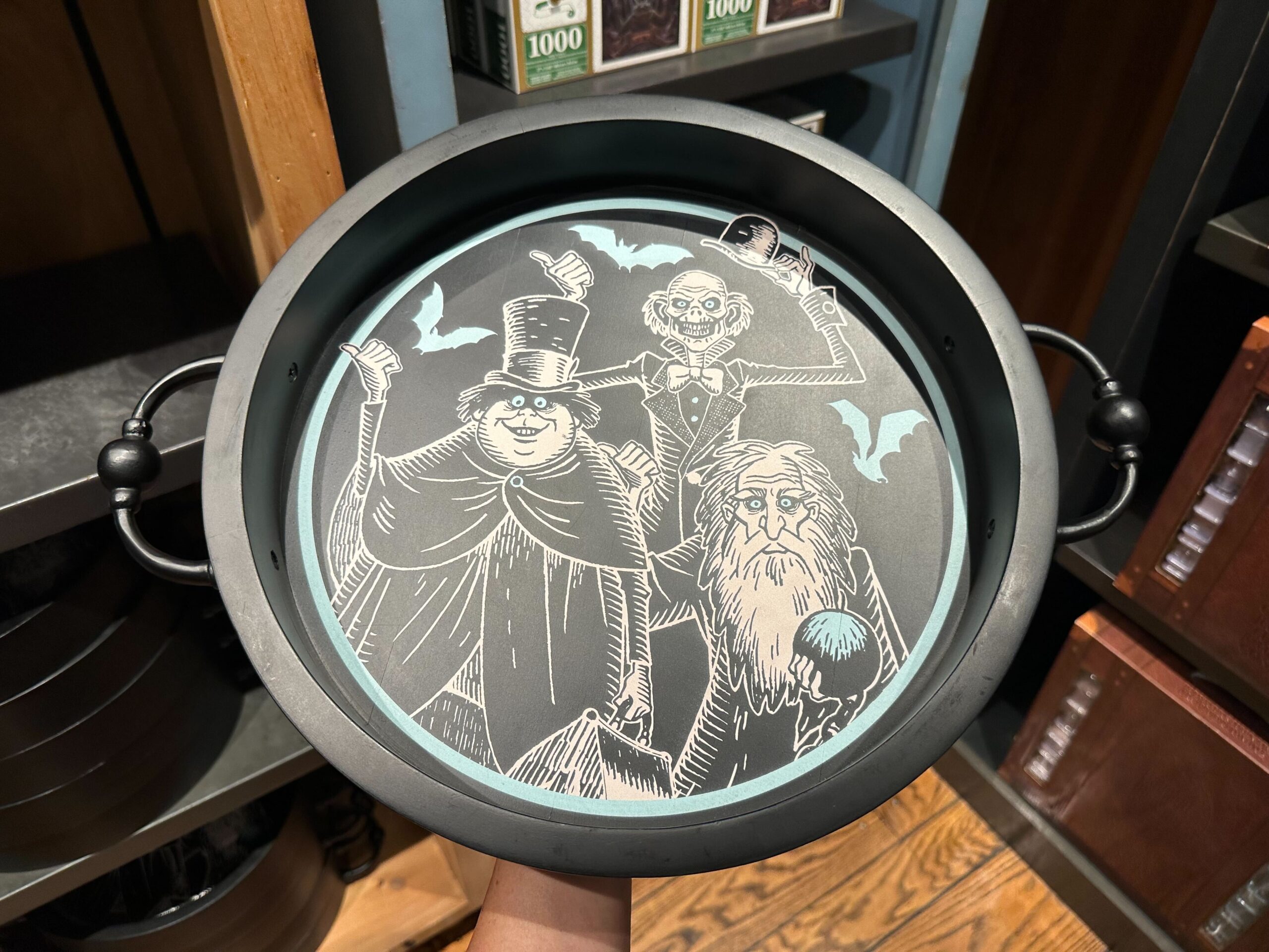 A round tray with black handles features three animated characters in Halloween costumes: a vampire, a ghost, and a bearded wizard. This fun piece of Halloween merchandise is perfect for adding some spooky charm to your decor.