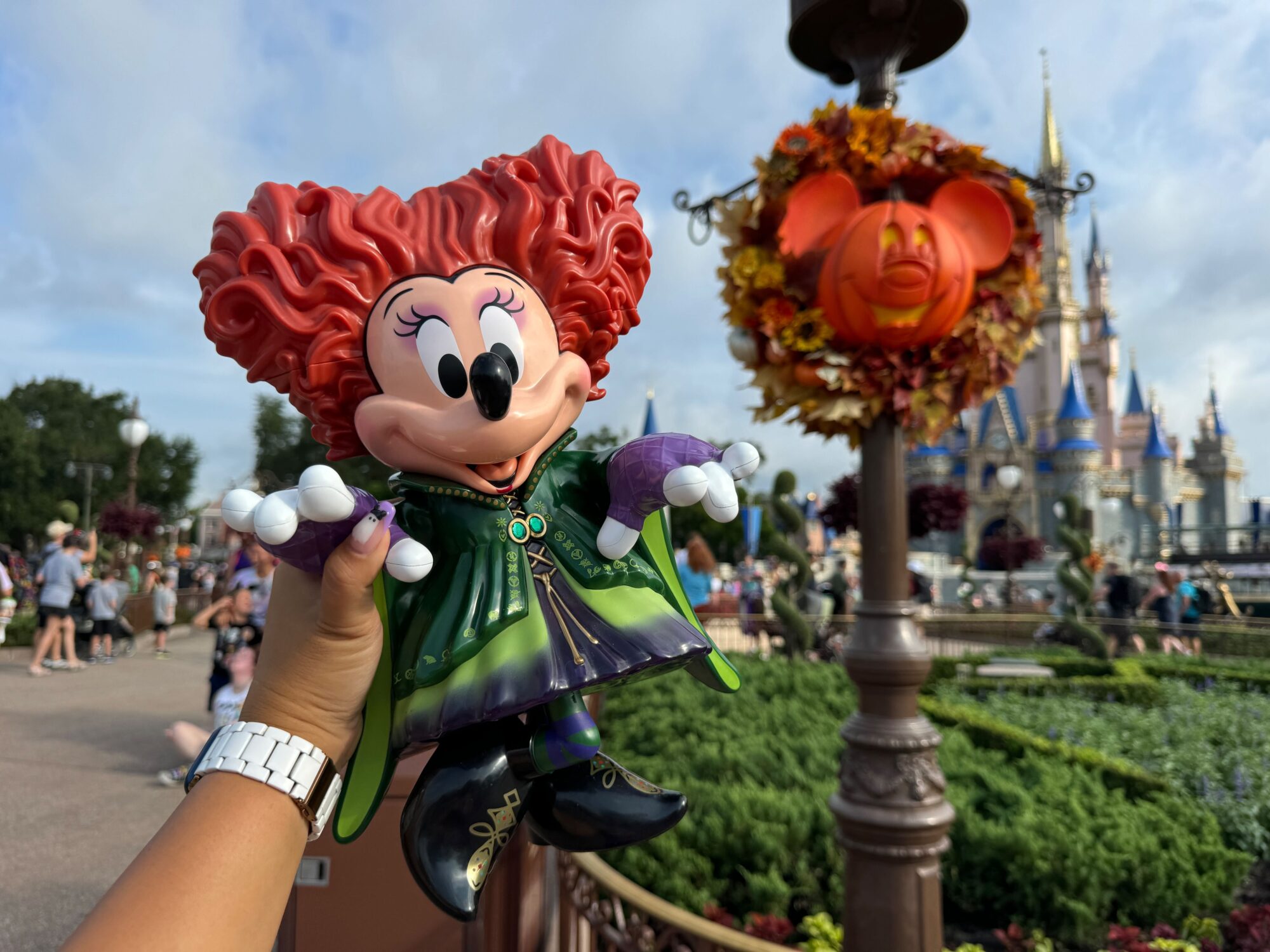 A hand holding a figurine of a red-haired character in a green dress and purple gloves, with a decorated Mickey Mouse face and castle in the background at a theme park.