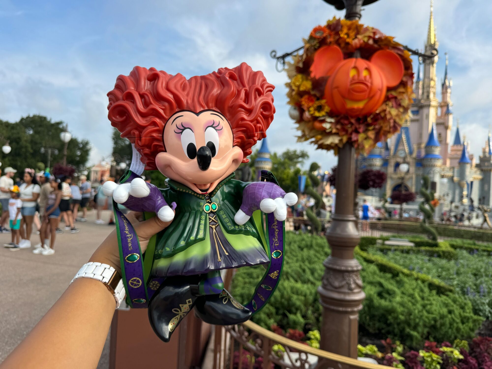 A hand holds a Minnie Mouse doll in a Halloween costume in front of a castle. The scene includes people, greenery, and a lamppost decorated with an orange Mickey Mouse pumpkin.