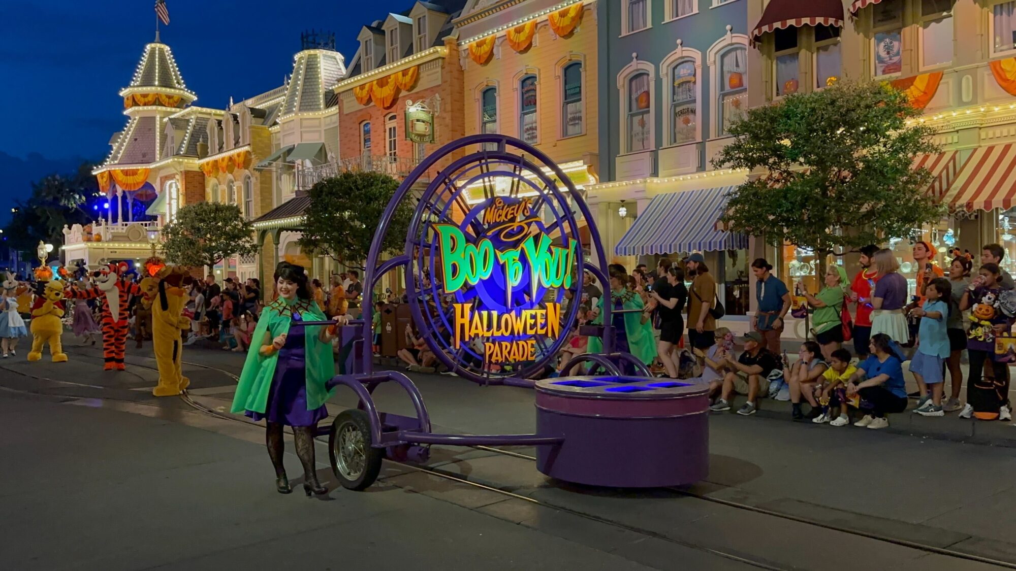 A person in a green and purple costume is walking alongside a purple parade float with a "Mickey's Boo to You Halloween Parade" sign, on a street decorated for Halloween, with spectators watching the exciting halloween parade.