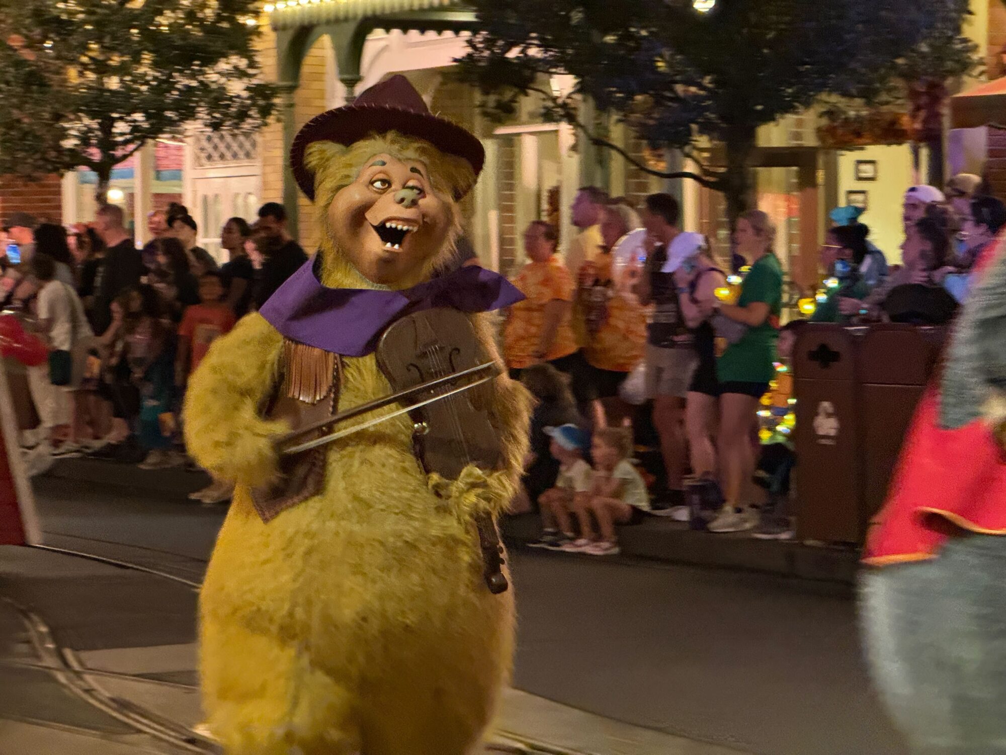 A person in a bear costume holding a fiddle walks in a Halloween parade at night, surrounded by spectators on a busy street decorated with lights.